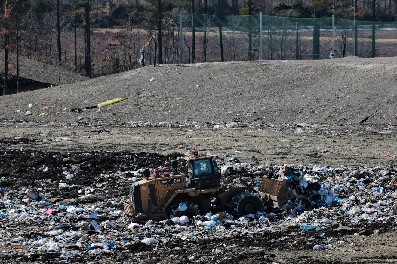 Bulldozer working amid scattered trash and debris at a landfill site with trees in the background.