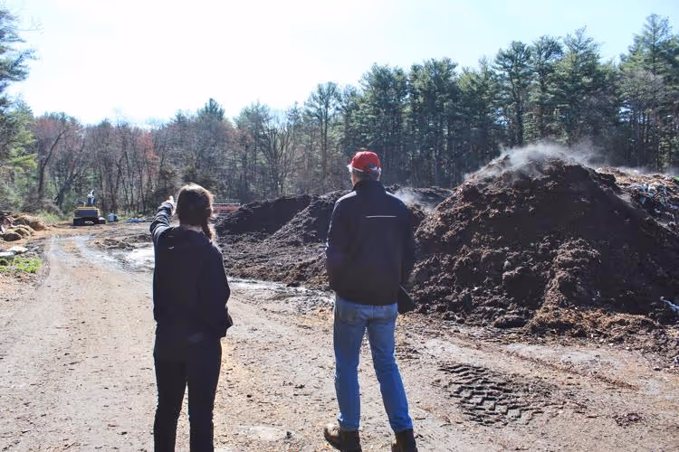 Two people standing on a dirt path near large steaming compost piles with trees in the background.
