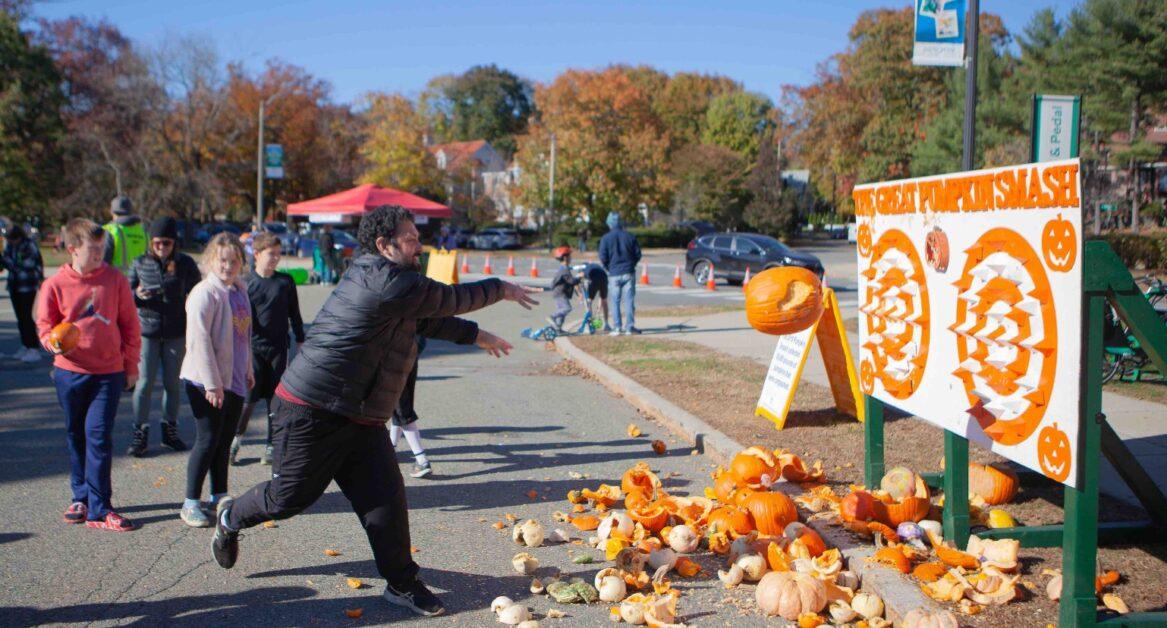 Group of people participating in a pumpkin smash game where pumpkins are thrown at a target board with pumpkin-shaped cutouts.