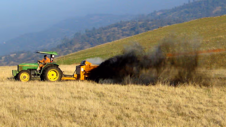 Green tractor pulling a machine that spreads dark compost material onto a dry grassy field with hills in the background.