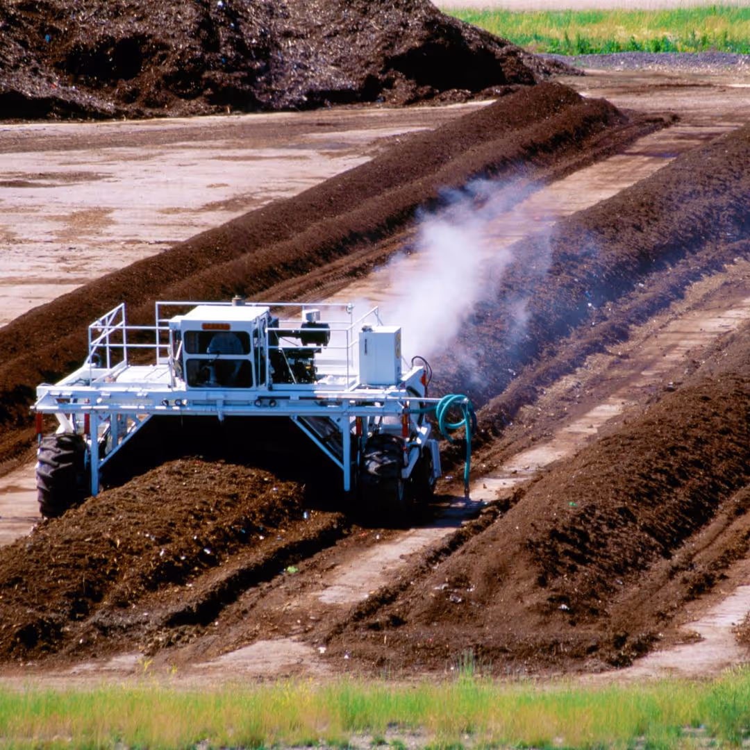 Large white machine turning and aerating rows of compost with steam rising from the organic material.