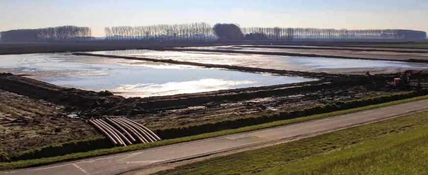 Large rectangular water treatment lined ponds with several exposed pipes crossing a road in the foreground, and a row of trees in the distance under clear sky.