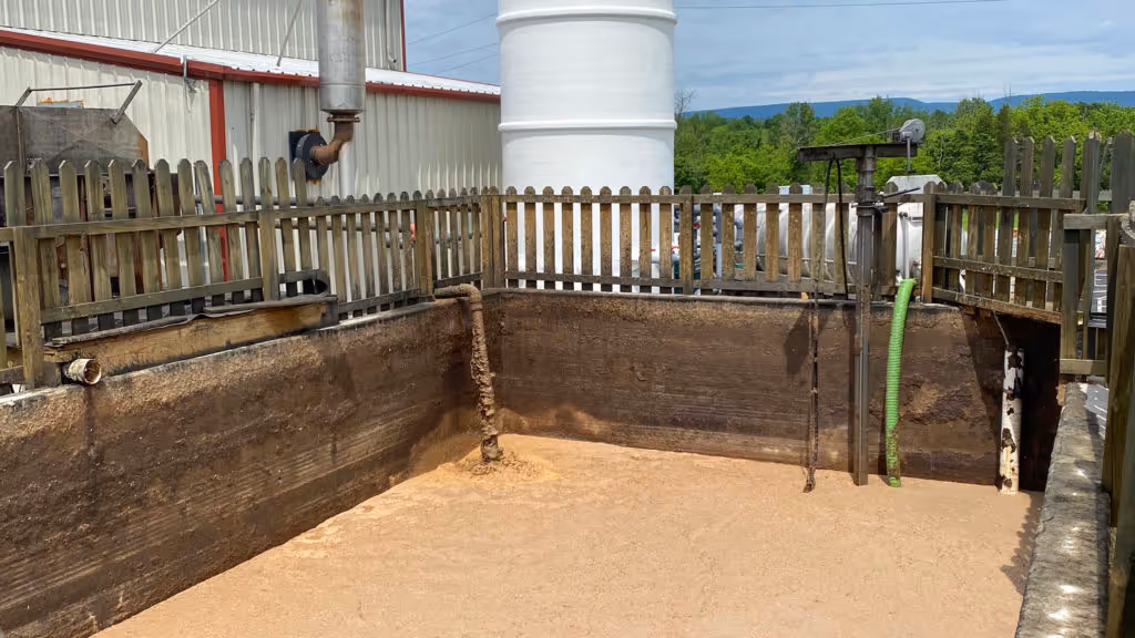 Large outdoor industrial tank or pit with muddy brown liquid inside, surrounded by wooden fencing and industrial equipment pipes.