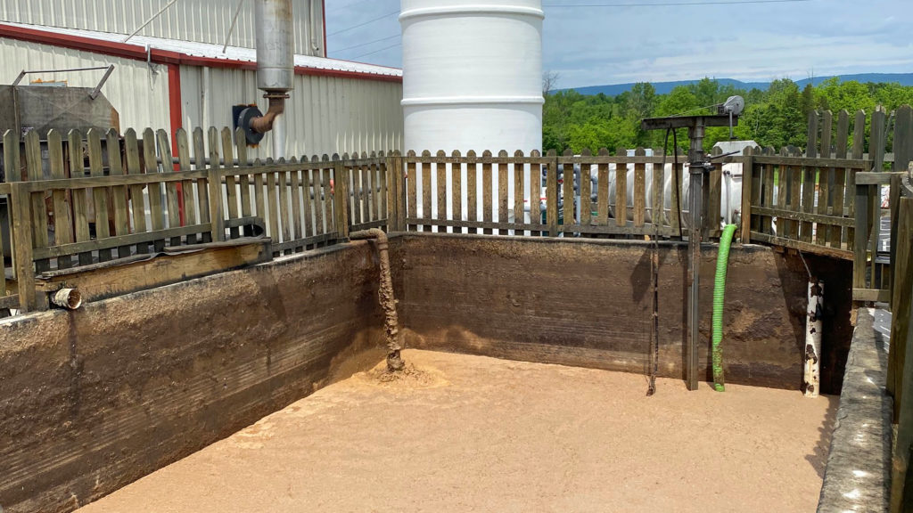 Large outdoor industrial tank or pit with muddy brown liquid inside, surrounded by wooden fencing and industrial equipment pipes.
