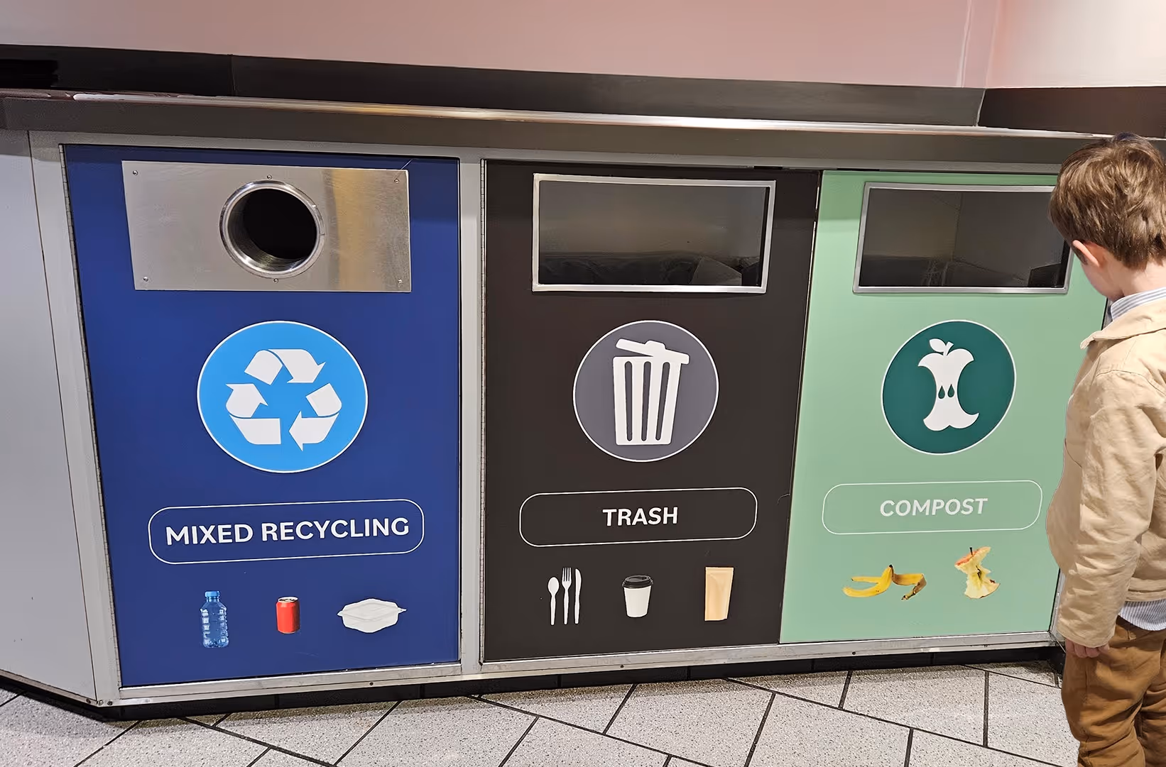 Three trash bins labeled Mixed Recycling with a bottle, can, and container, Trash with utensils, cup, and bag, and Compost with banana peel and apple core, with a child standing to the right.