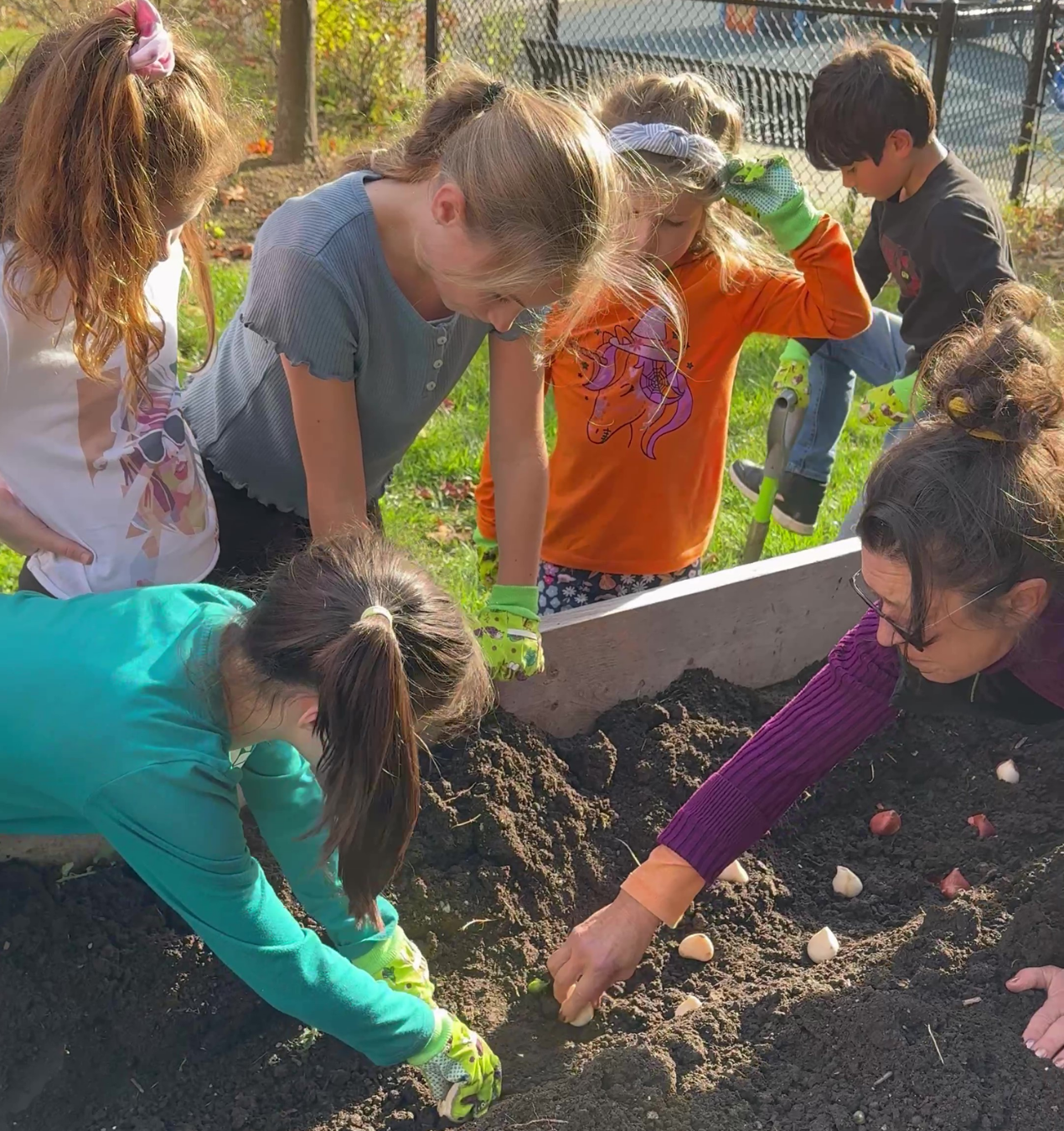 Group of children and an adult planting bulbs in garden soil outdoors.