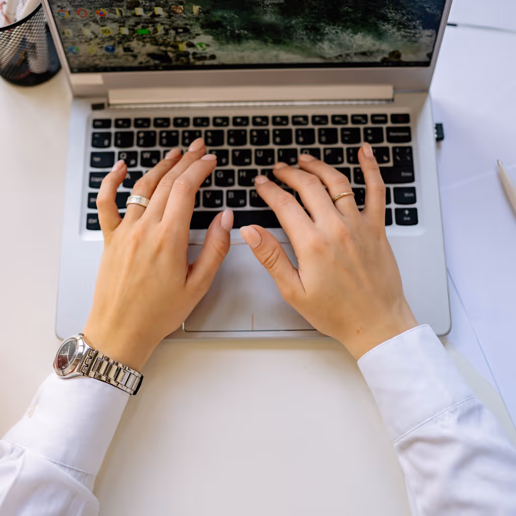 Person wearing a watch and rings typing on a laptop keyboard on a white desk.