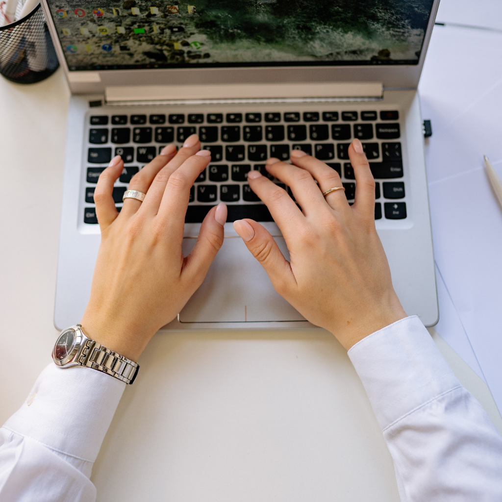 Person wearing a watch and rings typing on a laptop keyboard on a white desk.