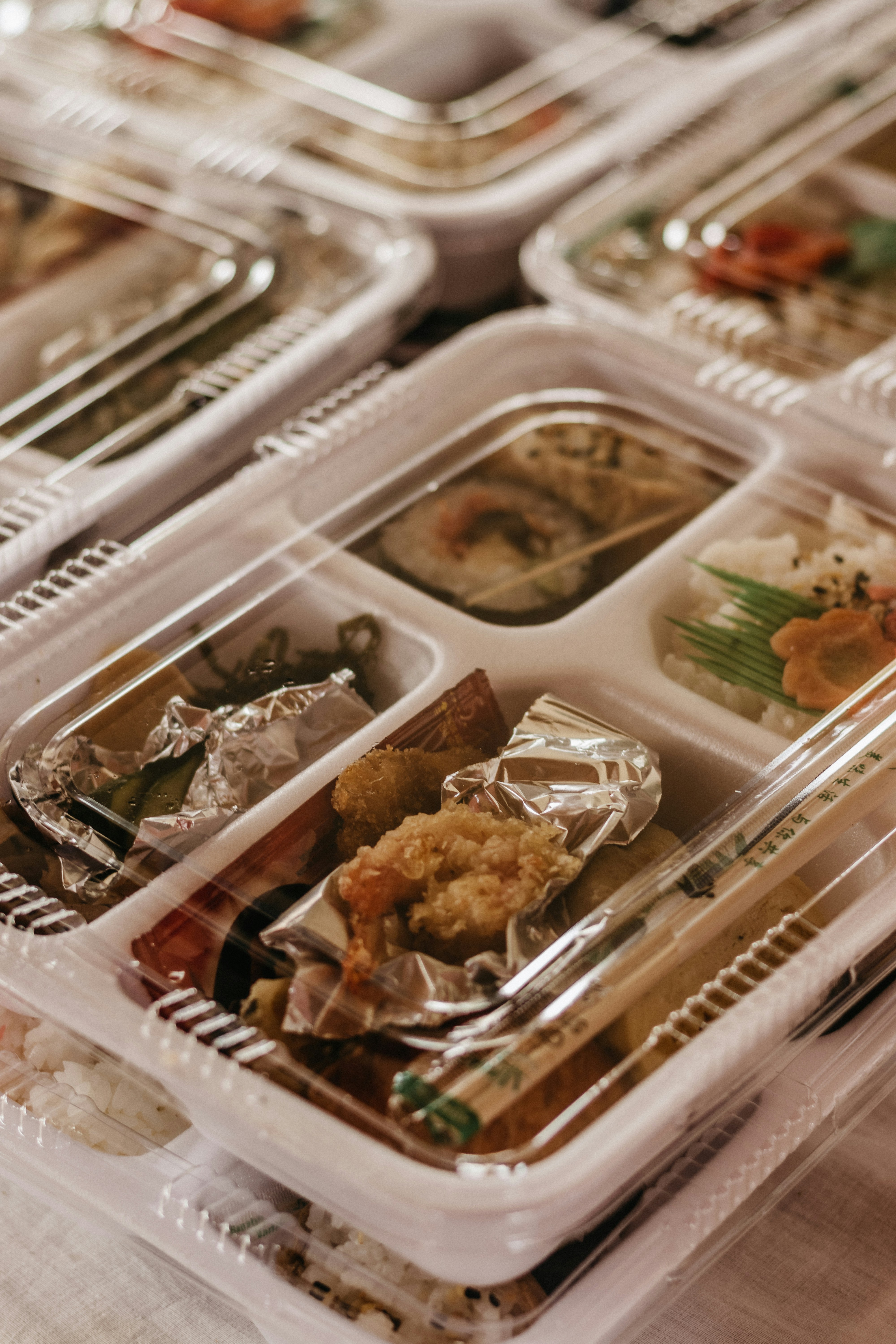 Plastic takeout container with compartments holding sushi, tempura shrimp, vegetables, rice, and chopsticks on top.