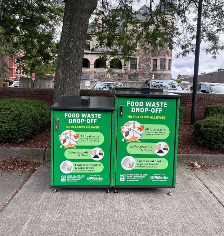 Two green food waste drop-off bins with instructions showing allowed items: all food waste, coffee grounds and filters, and food-soiled napkins and paper towels. No plastics allowed. Located outdoors on a sidewalk with trees and buildings in the background.
