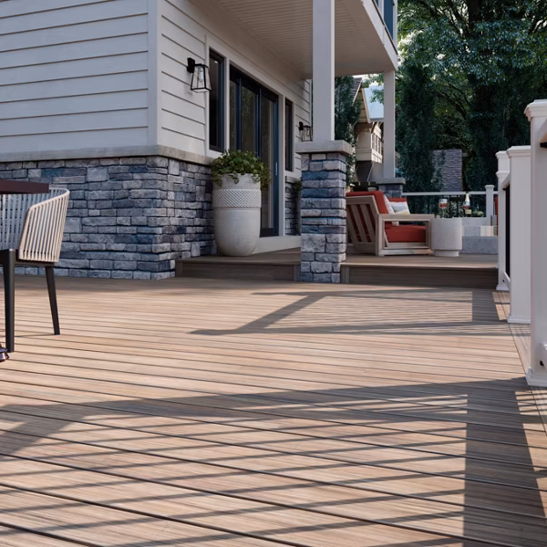 Spacious outdoor wooden deck attached to a house with stone siding, featuring patio furniture and shaded by trees.