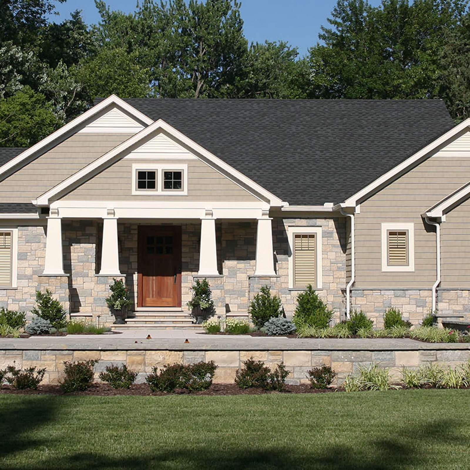 Modern single-story house with beige siding, stone facade, columns on front porch, and wooden front door surrounded by greenery.