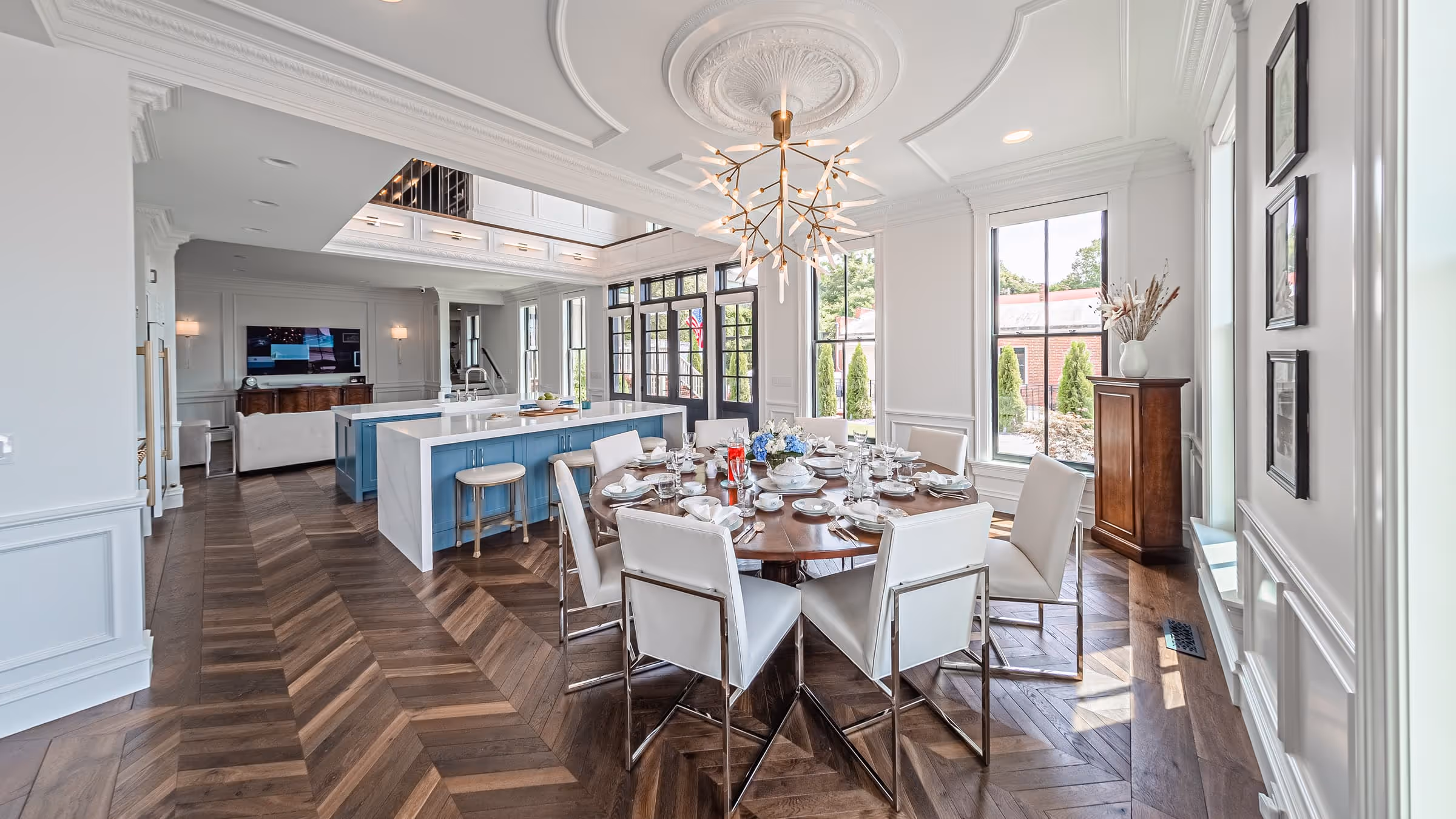 Spacious modern dining area with a round wooden table set for eight, white chairs, blue kitchen island, and large windows letting in natural light.