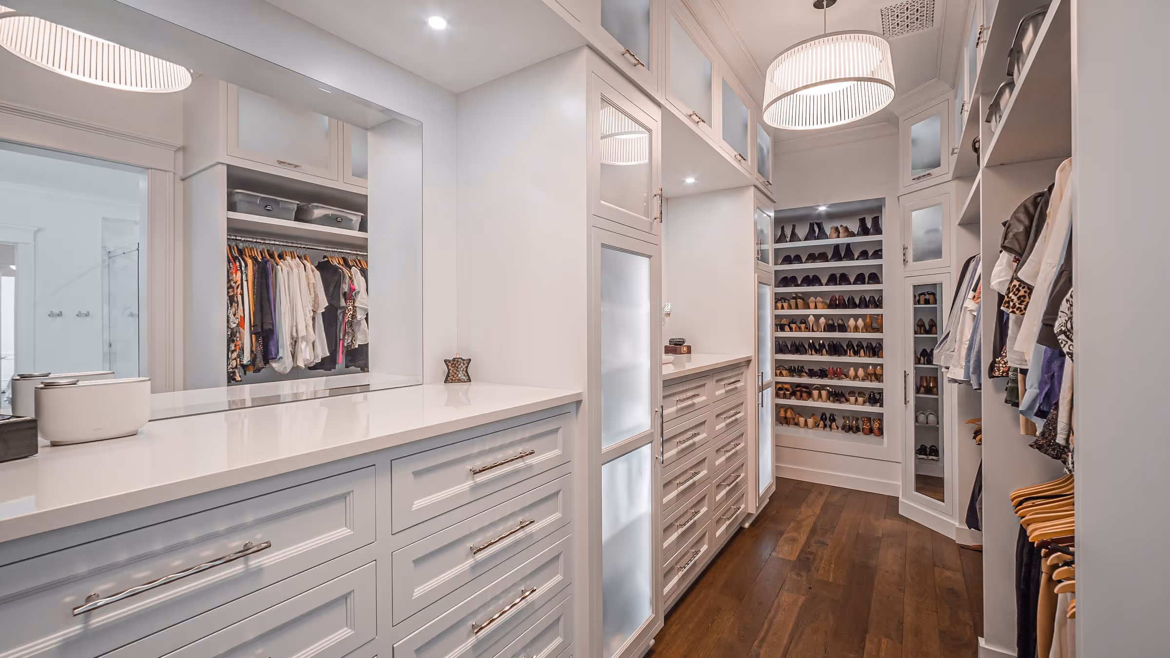 Modern walk-in closet with white cabinetry, hanging clothes, built-in shoe shelves, and wooden flooring.