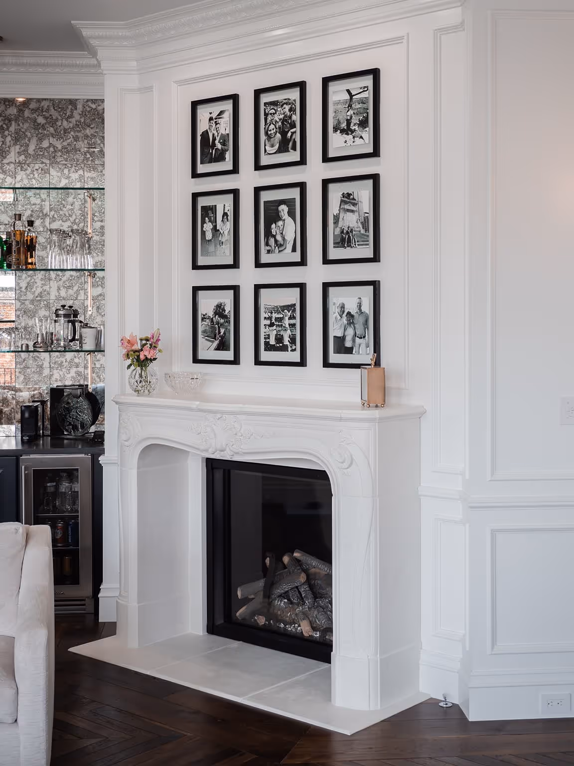 White ornate fireplace with nine black-framed black-and-white family photos arranged in a 3x3 grid above it, next to a mirrored bar with glass shelves and a beige sofa on dark hardwood floors.