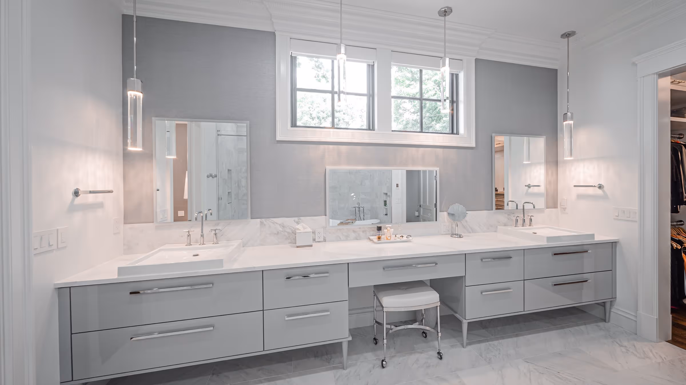 Modern bathroom with double sinks, light gray cabinets, three mirrors, hanging pendant lights, and a small cushioned stool on wheels.