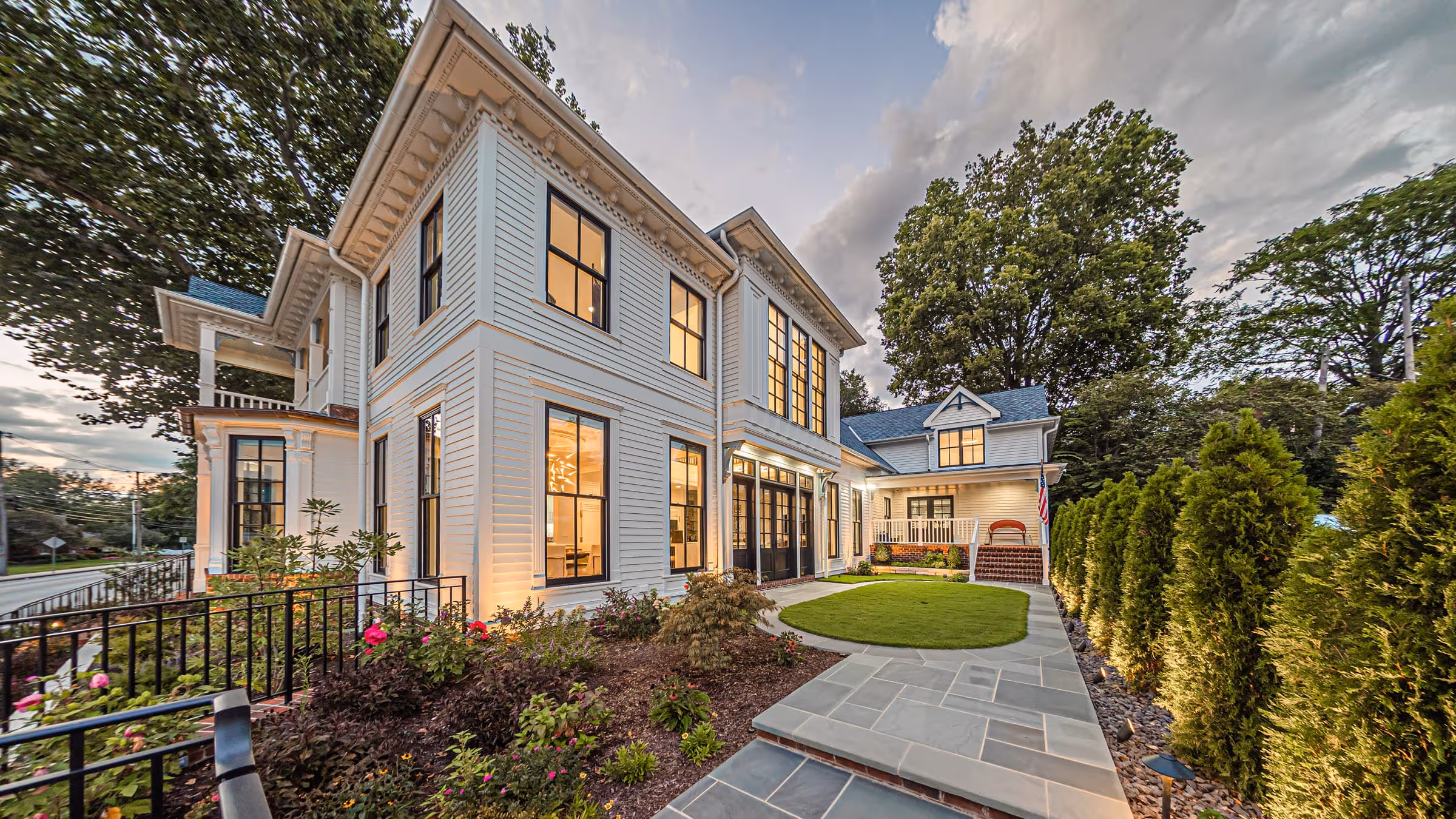 Large modern Victorian-style white house with black window frames, lit interior, landscaped garden, and paved walkway.
