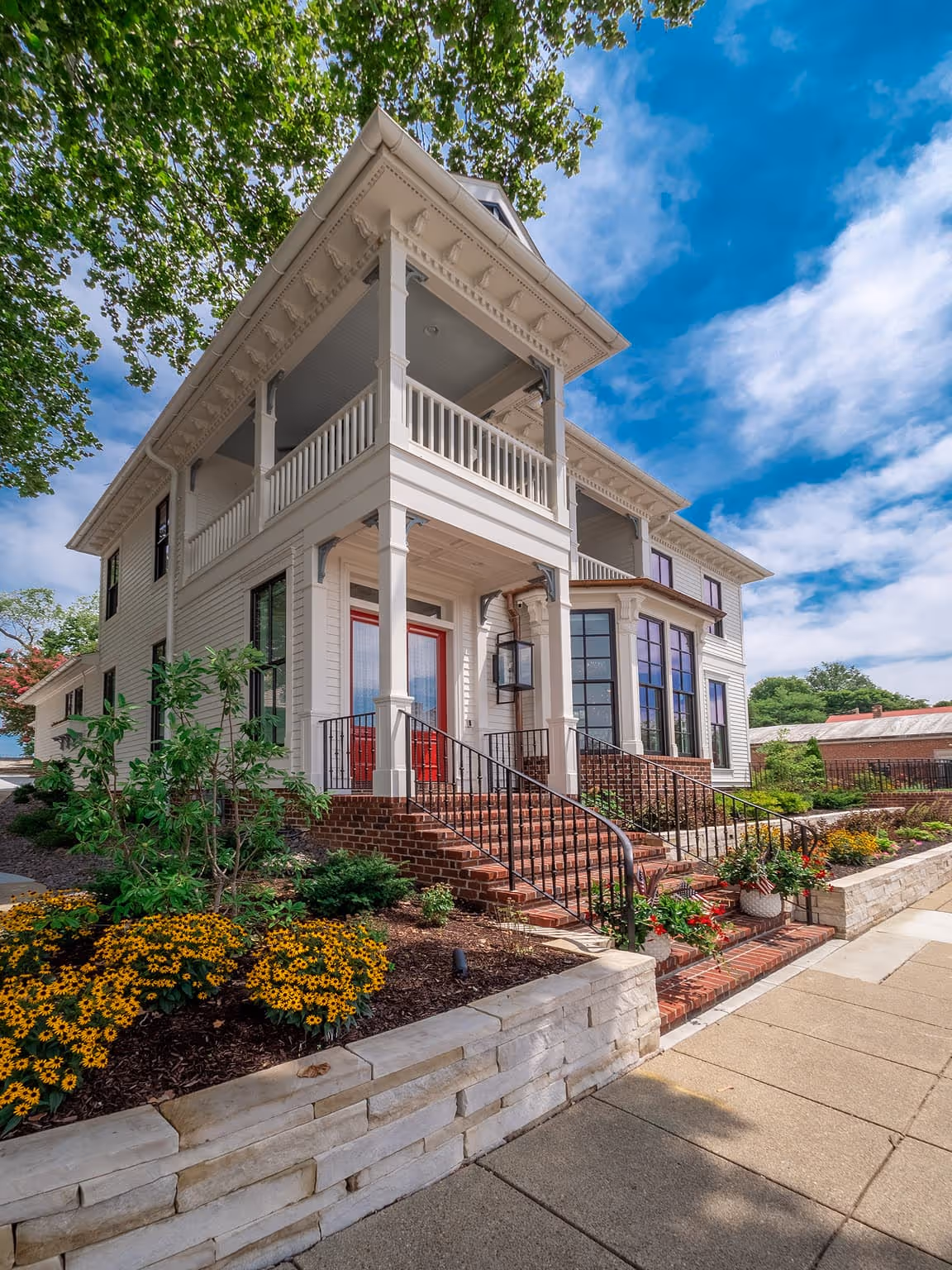White modern Victorian-style house with red double doors, brick steps, and a landscaped garden under a blue sky.