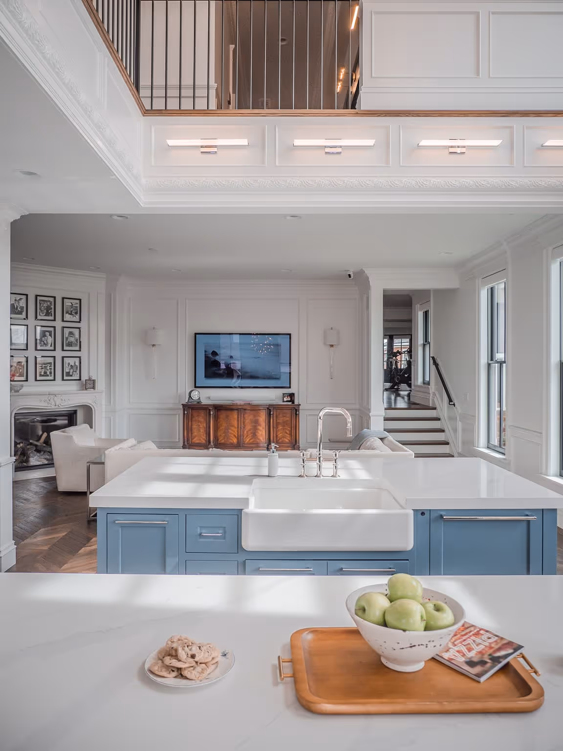 Modern kitchen with blue island, white farmhouse sink, bowl of green apples, plate of cookies, and a living room with wall-mounted TV in the background.