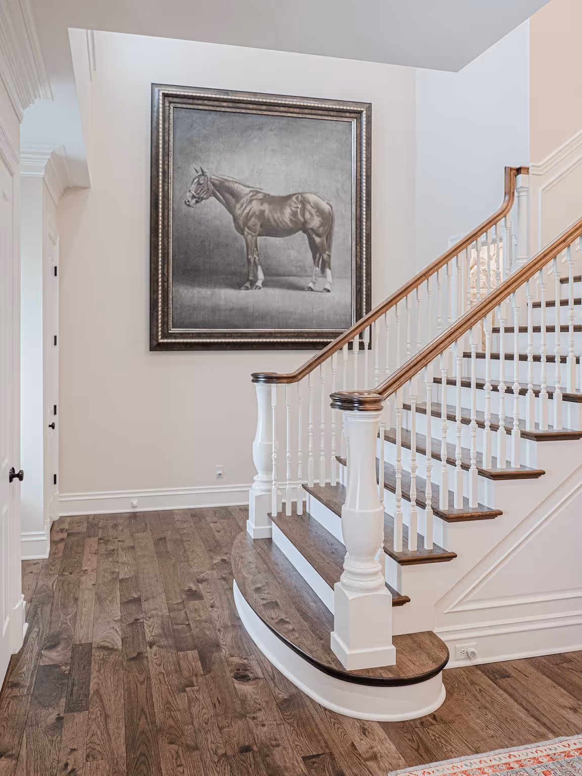 Interior view of a staircase with wooden steps and white balusters, next to a wall with a large framed grayscale horse painting.