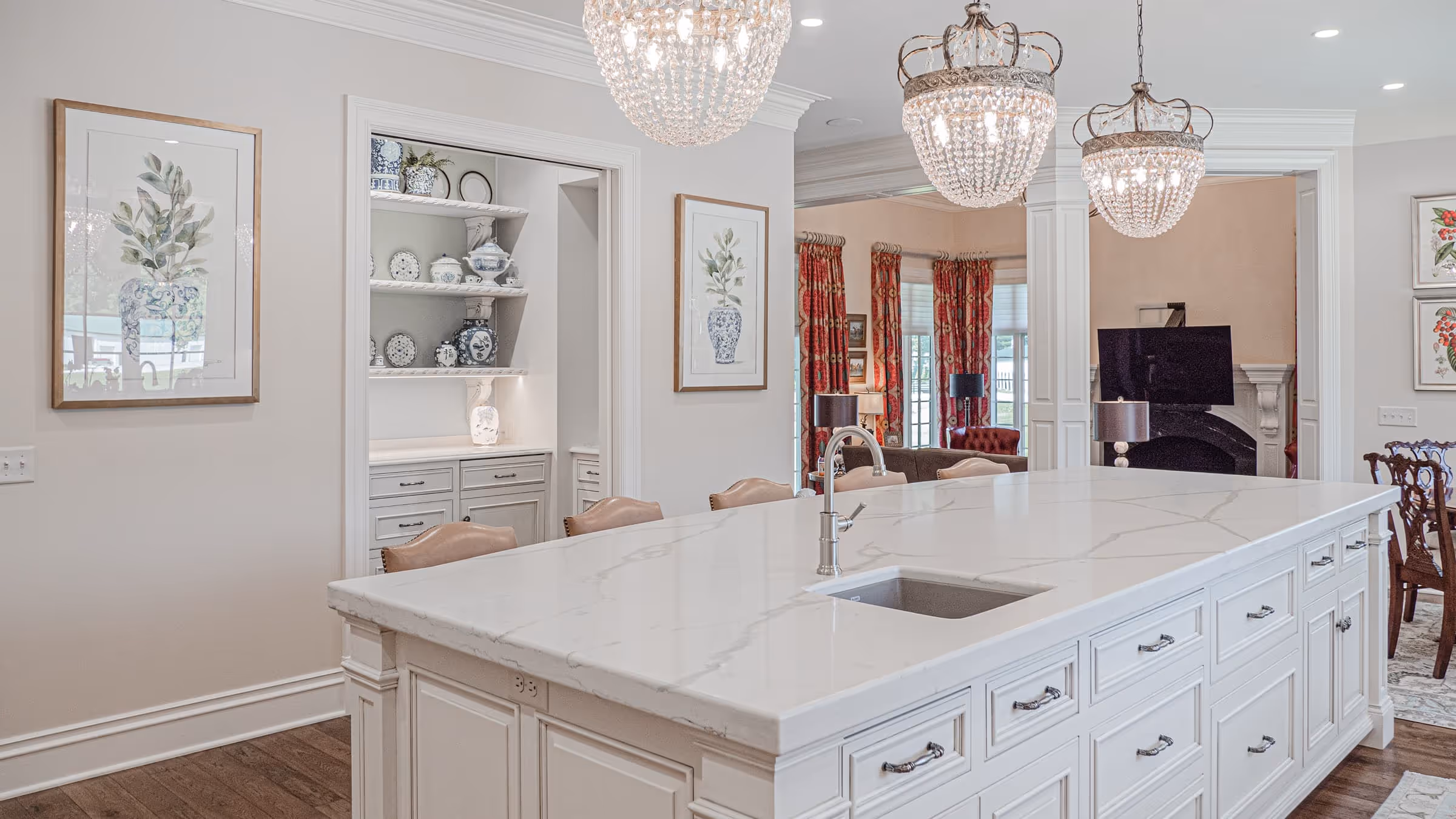 Spacious kitchen with a large white marble island featuring a built-in sink and leather bar stools, crystal chandeliers overhead, and decorative shelves with blue and white ceramics.