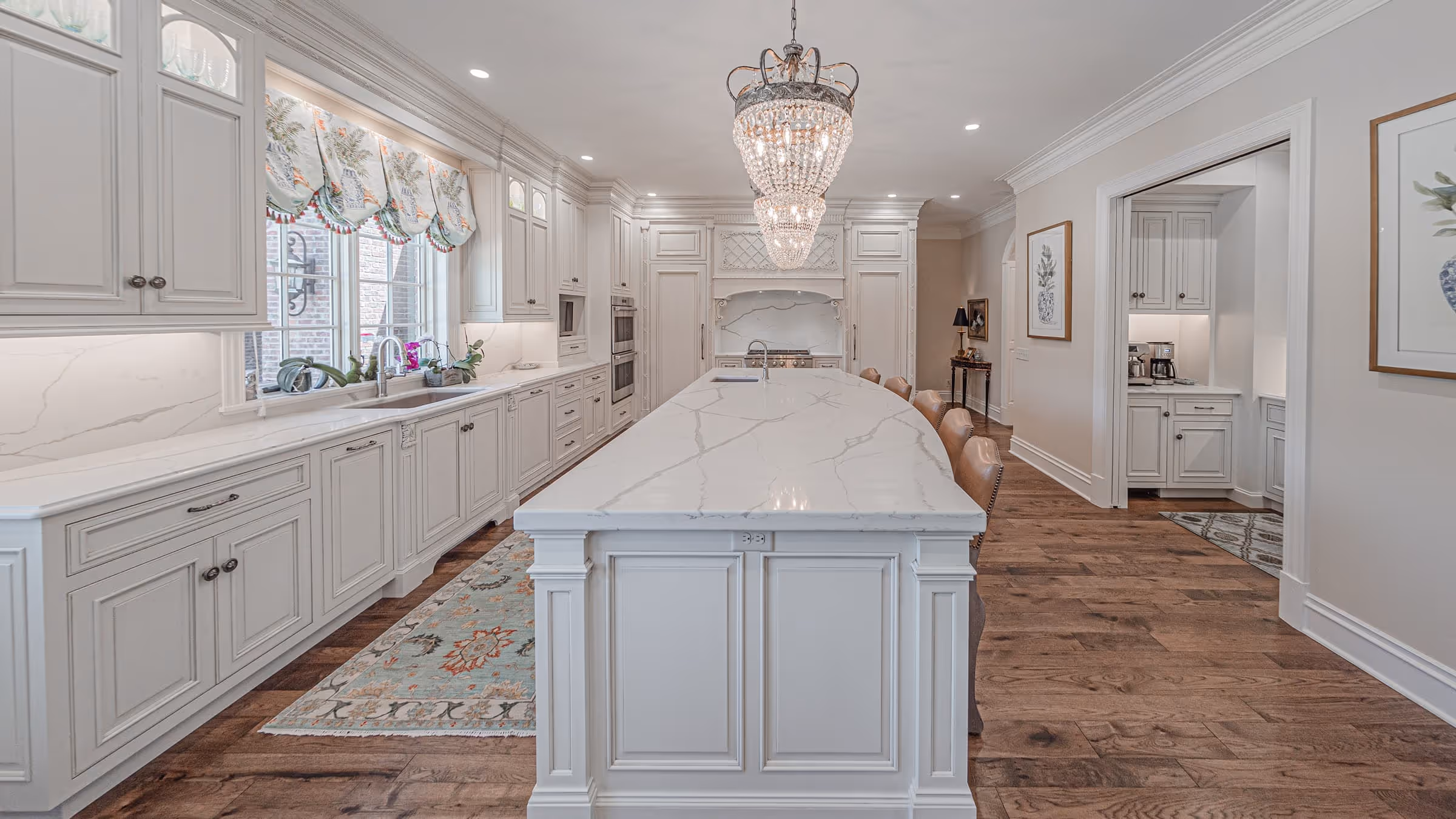 Spacious traditional kitchen with white cabinetry, a large marble island, crystal chandeliers, hardwood floors, and decorative rugs.
