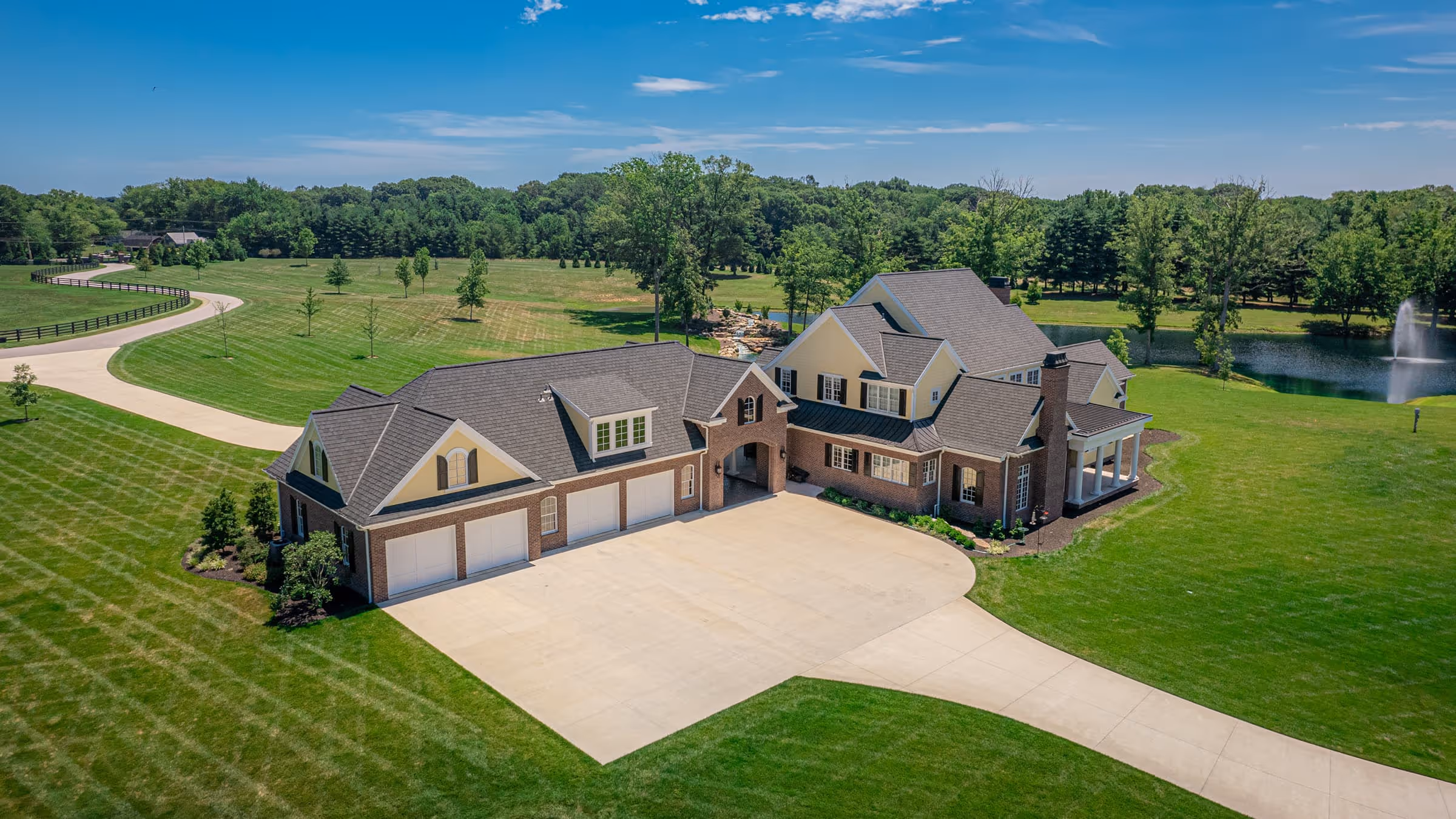 Traditional Southern house with multiple gables, four-car garage, spacious driveway, surrounded by green lawn and trees, with a pond and fountain in the background under a blue sky.