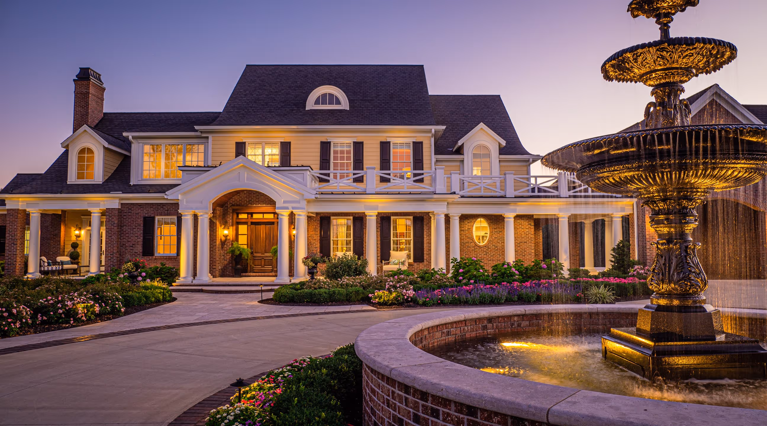 Elegant large tTraditional Southern house with lit windows, white columns, landscaped garden, and a decorative fountain in the foreground at dusk.