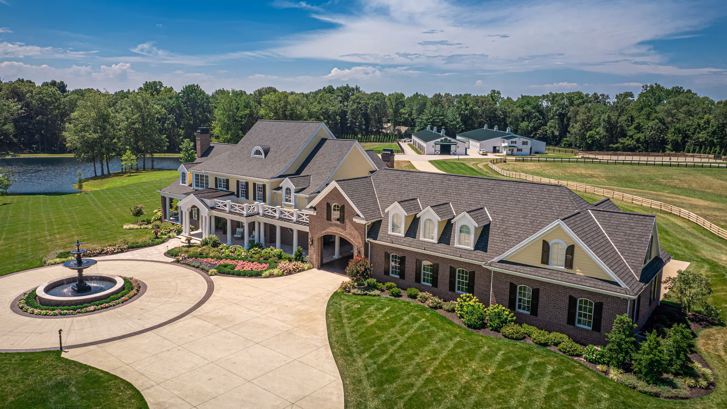 Large traditional southern estate with a circular driveway, central fountain, manicured gardens, and surrounding green lawn with trees and a pond.