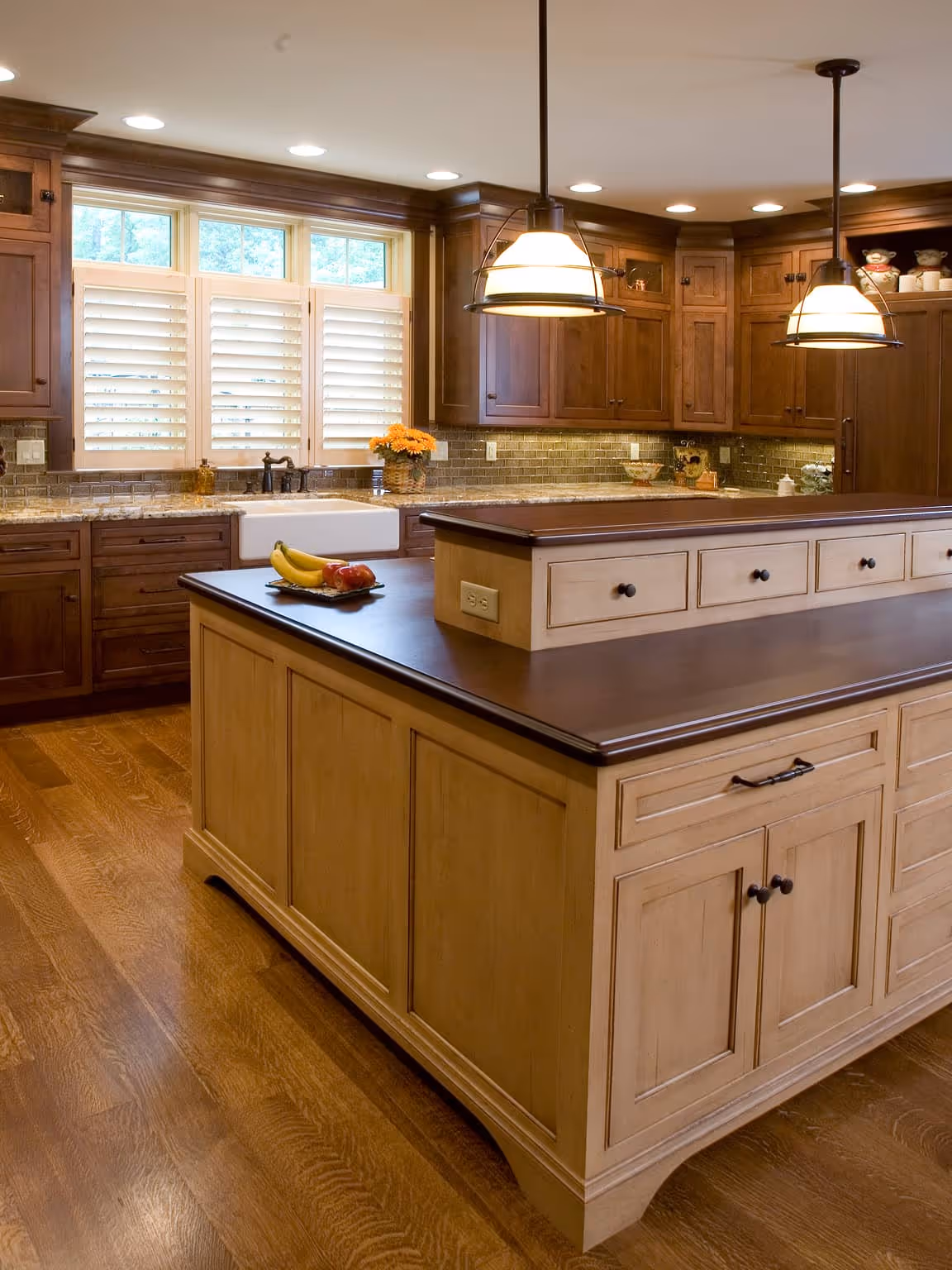 Traditional kitchen with a large wooden island, hanging pendant lights, dark wood cabinets, and a farmhouse sink under three shuttered windows.