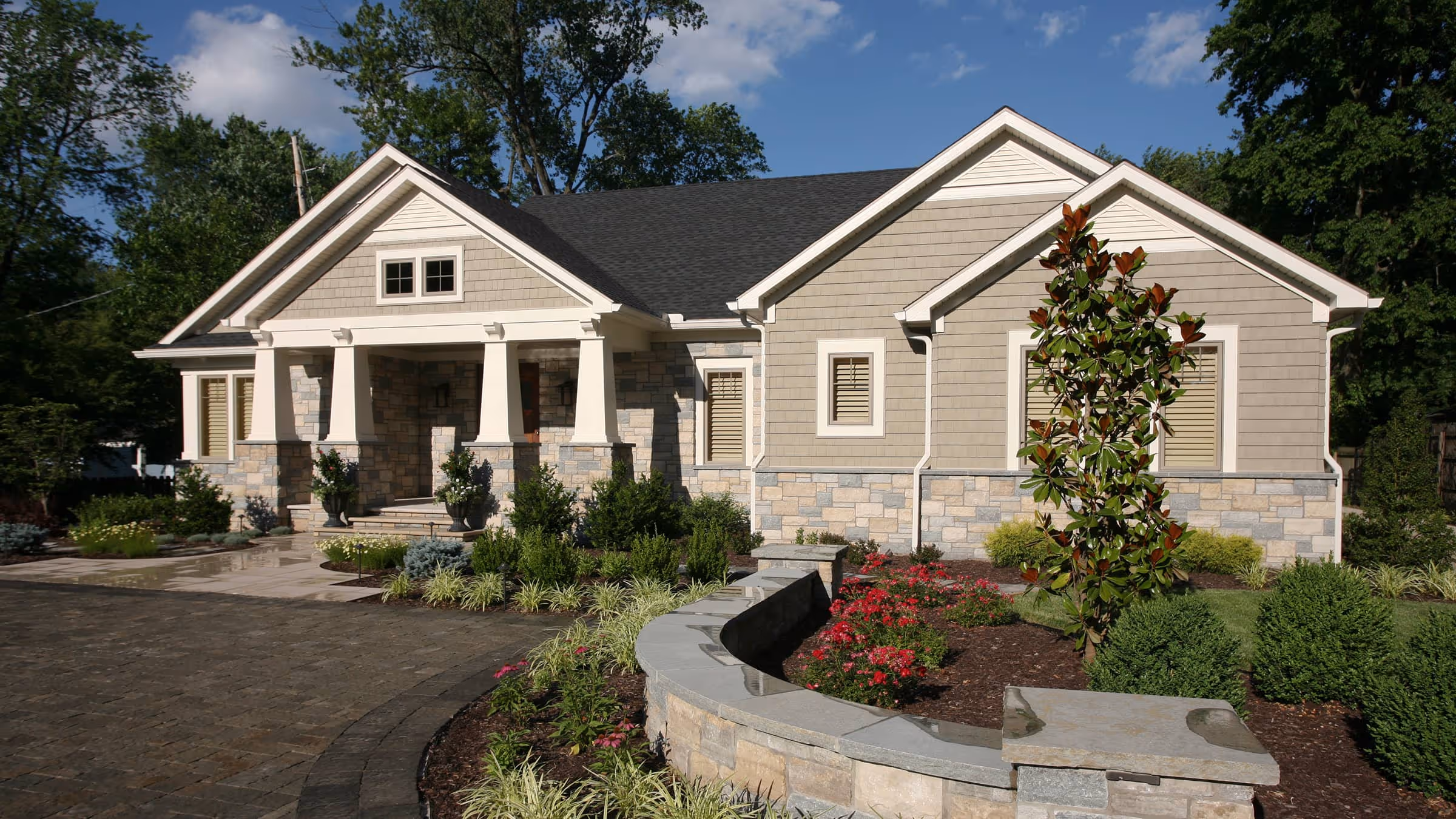 Single-story craftsman style house with stone and shingle exterior, front porch with white columns, and landscaped garden with curved stone wall and red flowers.