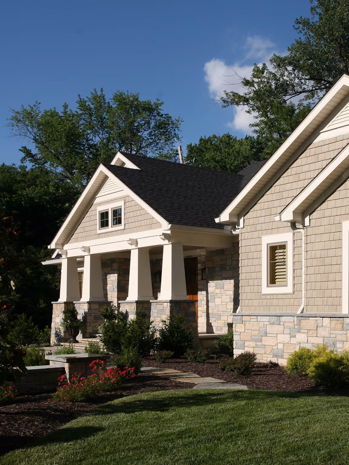 Craftsman-style house with beige siding, stone pillars, and a black roof under a clear blue sky.