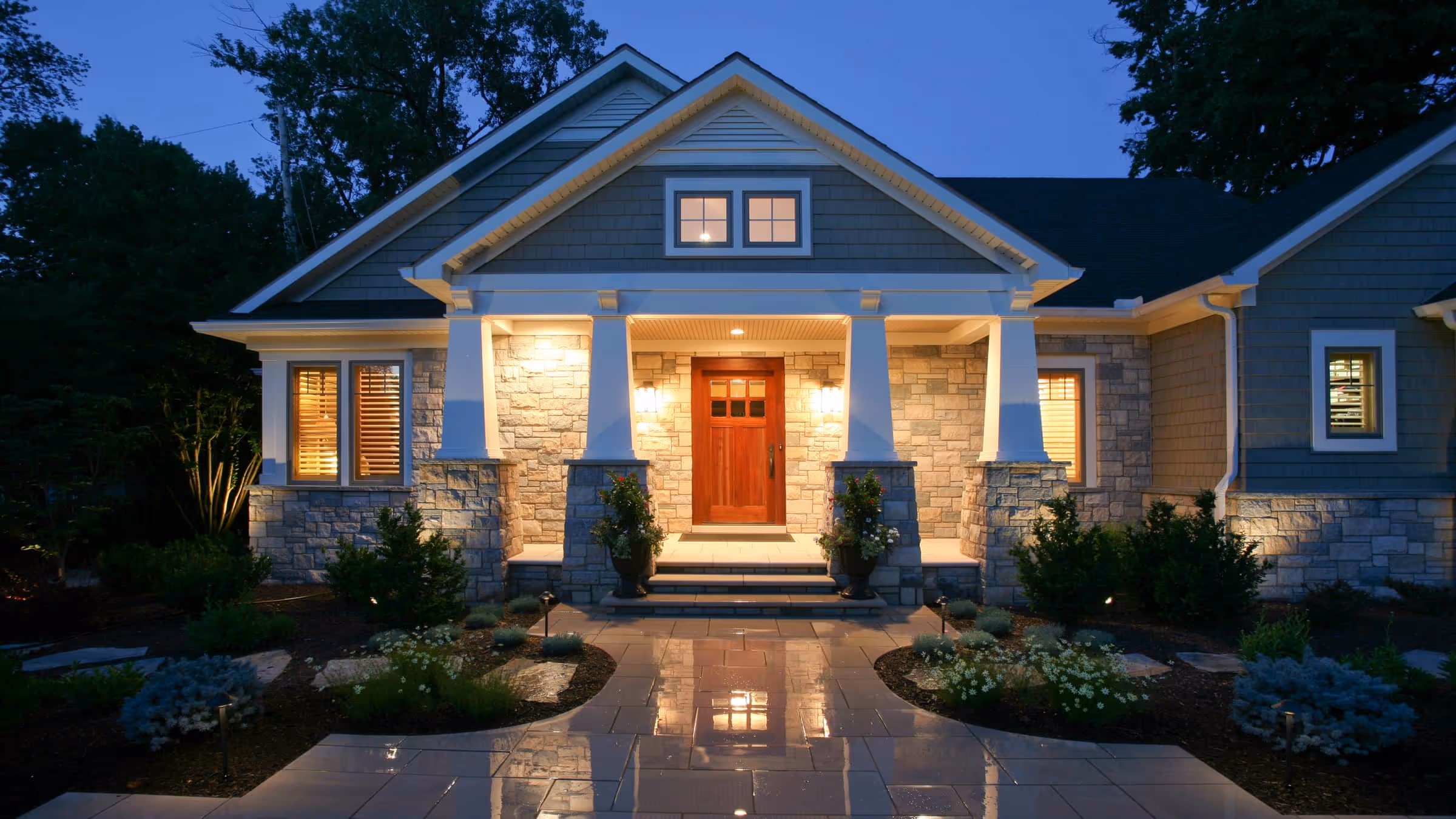 Craftsman-style house entrance at dusk with stone pillars, wooden front door, lit porch lights, and reflective stone pathway.