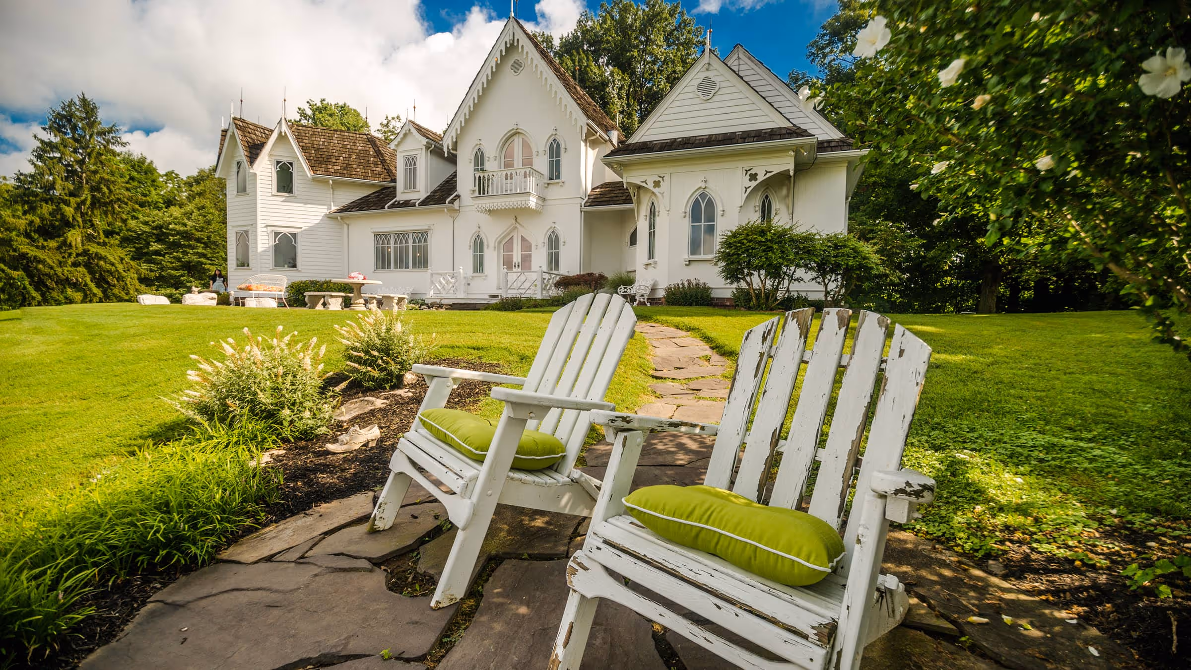 White Adirondack chairs with green cushions on a stone path leading to a large white Gothic Revival house surrounded by green lawn and trees.
