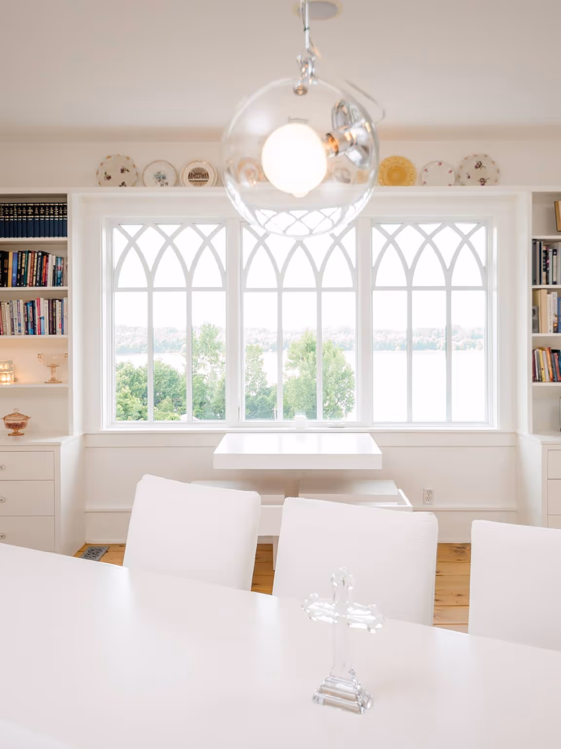 Bright dining area with white chairs, a white table featuring a glass cross, large gothic-style windows overlooking trees and water, and built-in bookshelves.