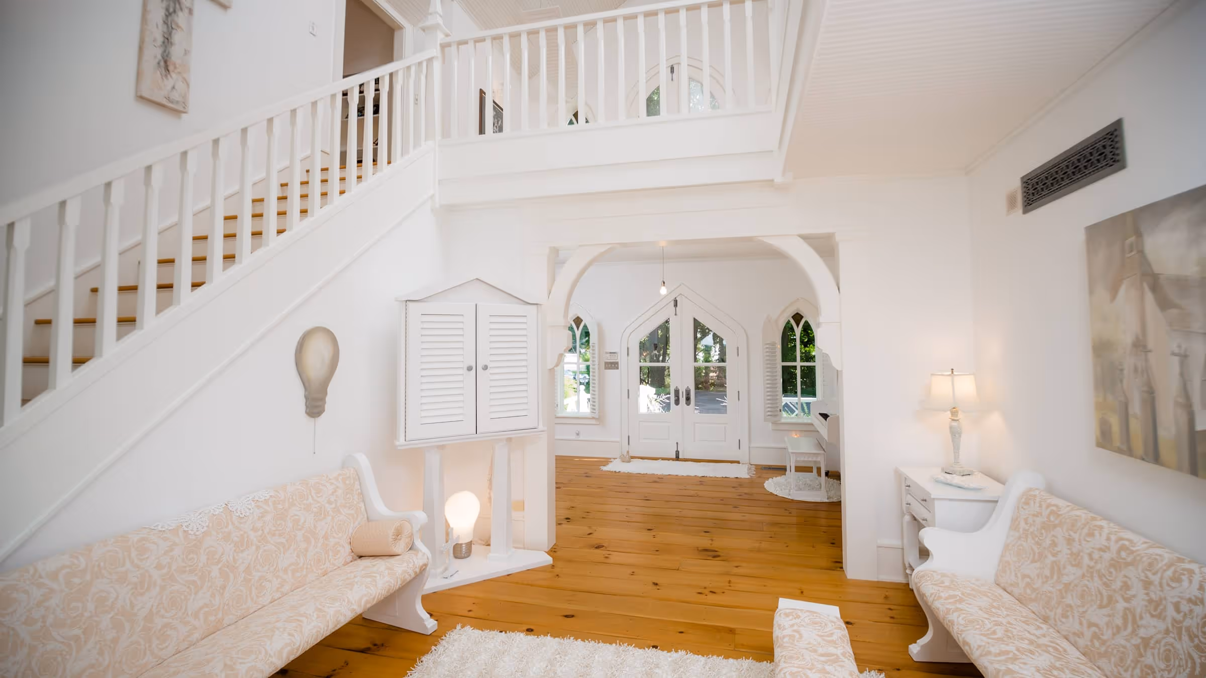 Bright white Gothic Revival interior with wooden floor, arched windows and doors, patterned benches, and staircase with white railing.