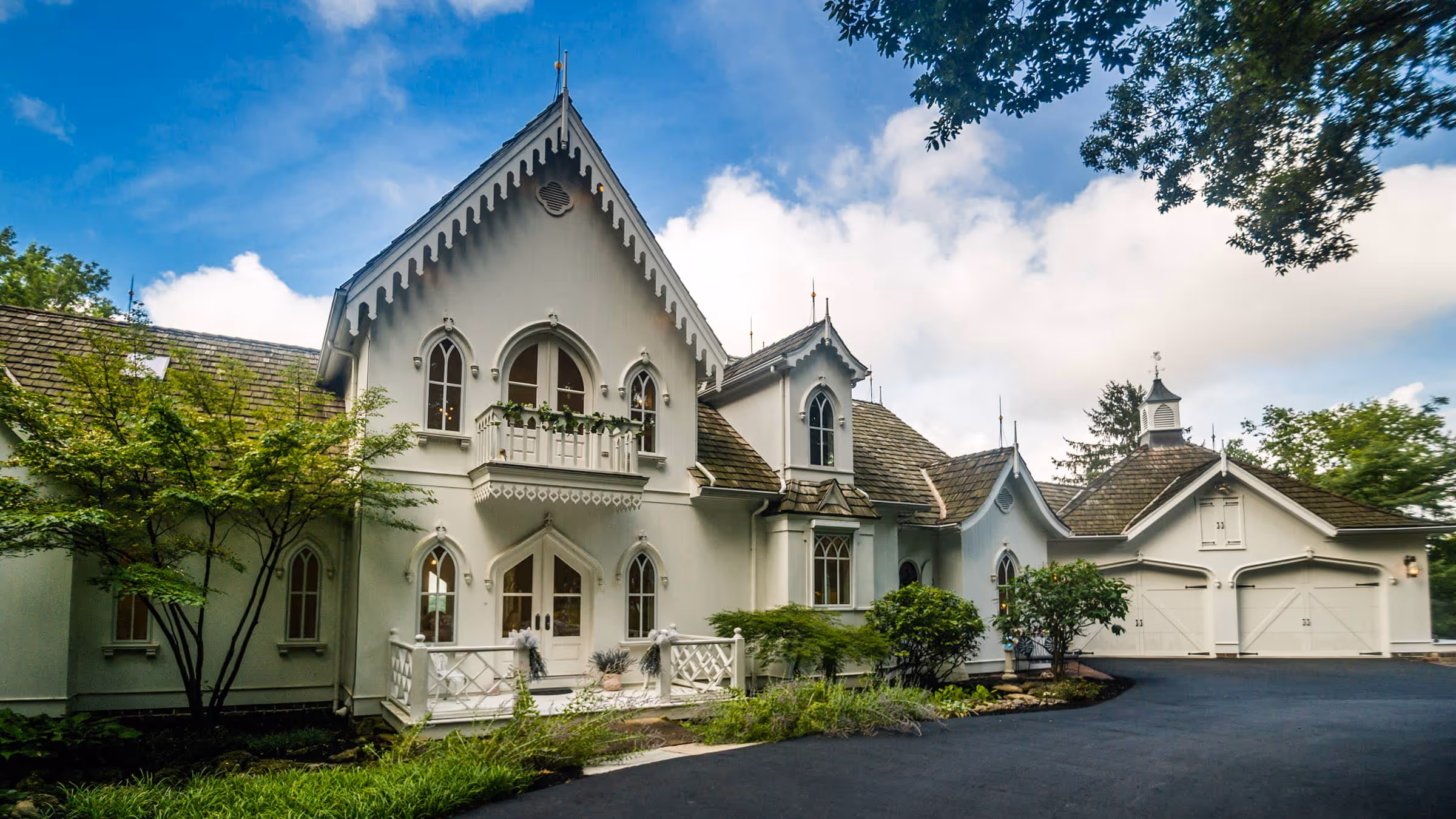 Large white Gothic Revival style house with pointed arches, balcony, and attached garage under a partly cloudy sky.