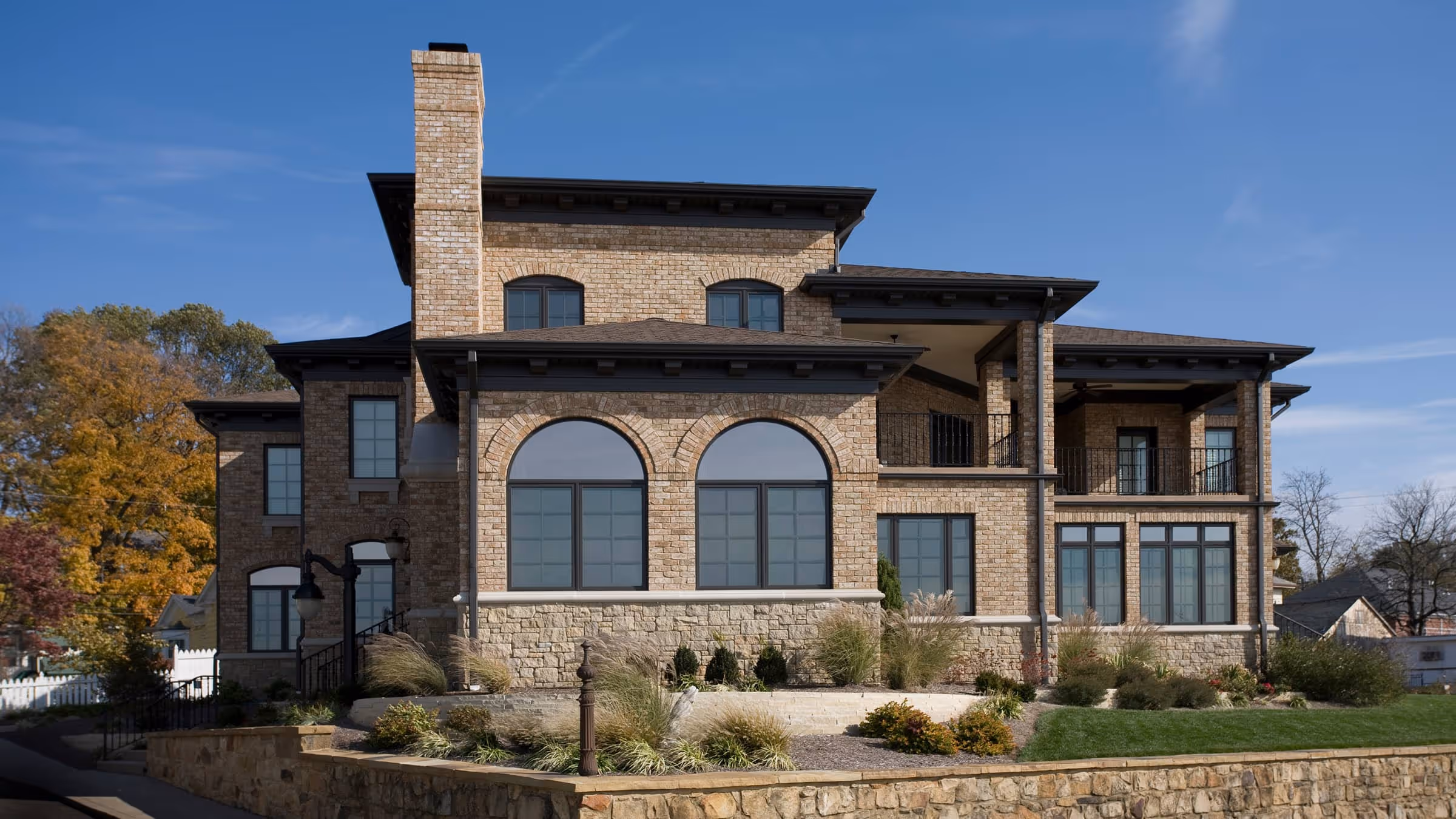 Large two-story brick California Mediterranean style house with arched windows, black trim, and a stone foundation bordered by landscaped bushes and grass under a blue sky.