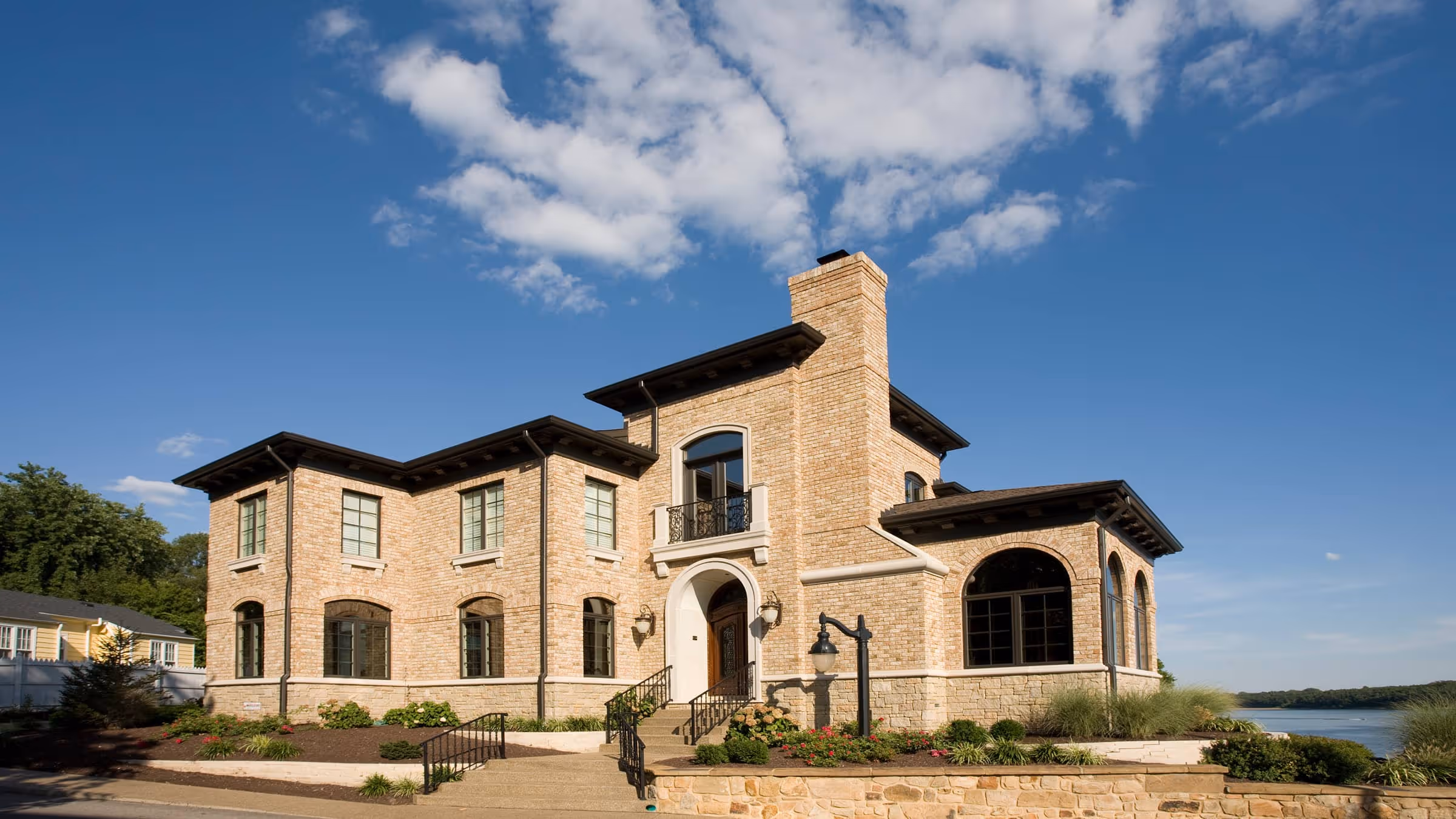 Two-story brick California Mediterranean style house with arched windows and a stone foundation under a partly cloudy blue sky.