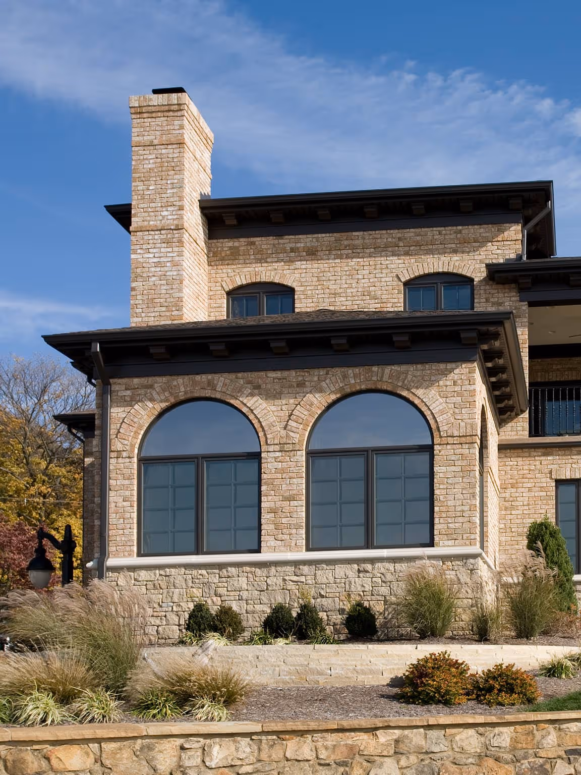 Large brick California Mediterranean style house with arched windows, stone foundation, and a brick chimney under a blue sky.