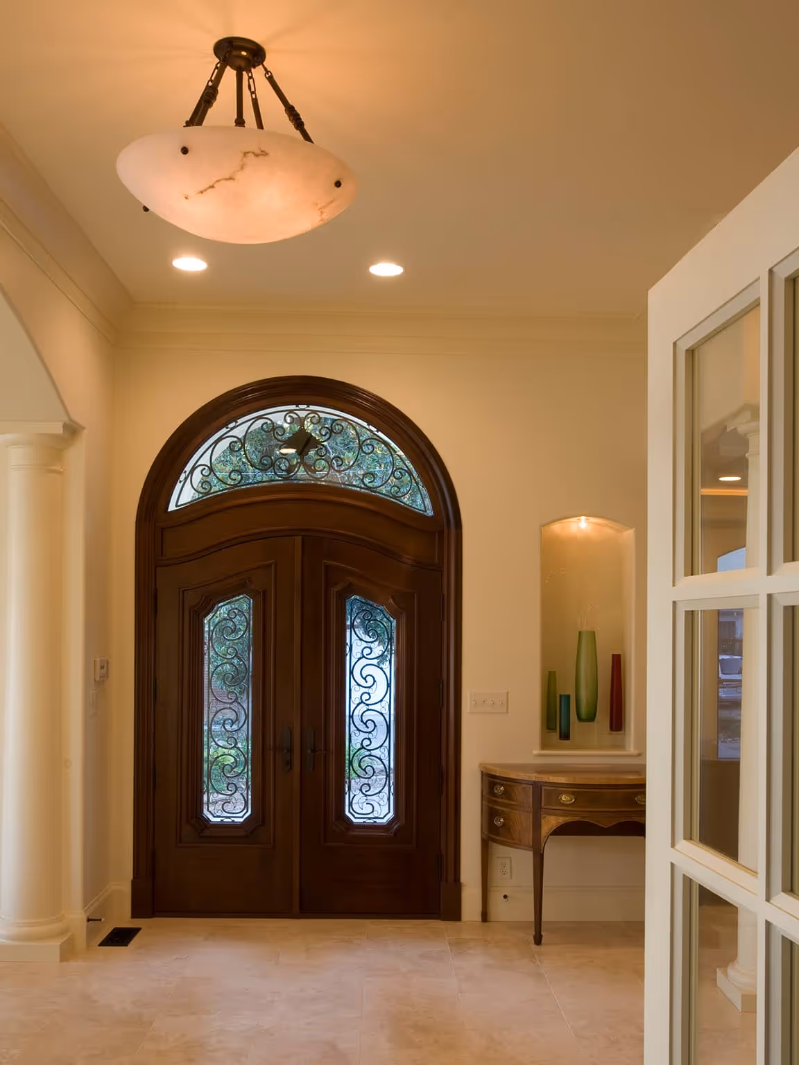 Entryway with double wooden arched doors featuring decorative wrought iron details, a vintage console table with glass vases in a recessed niche, and a ceiling light fixture.