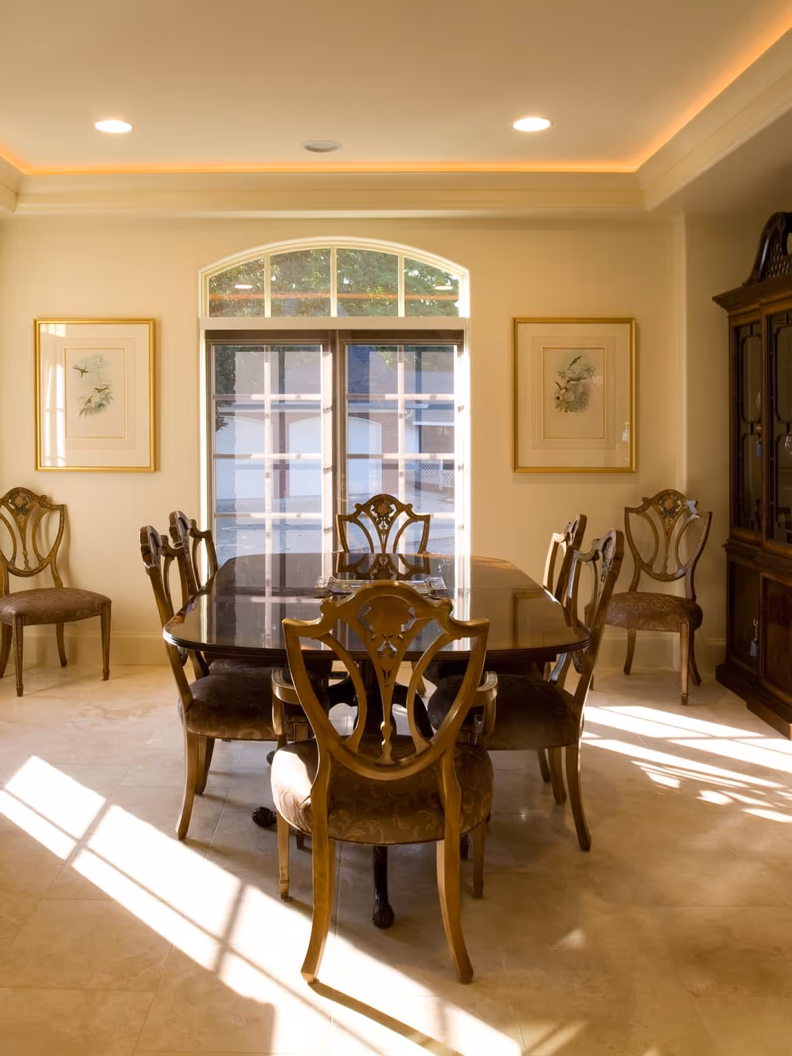 Elegant dining room with a polished wooden table, eight intricately carved chairs, two framed botanical prints, and sunlight streaming through a window.