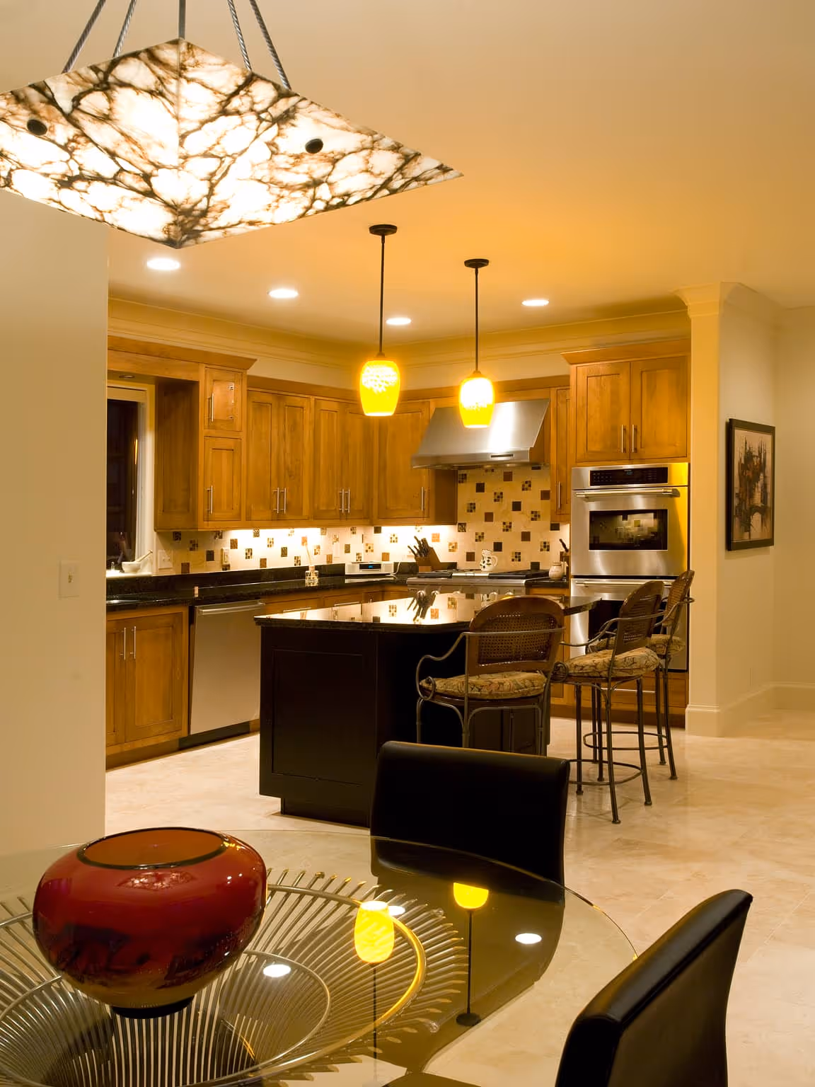 Warmly lit kitchen with wooden cabinets, a black island with three cushioned stools, and a glass dining table with a red vase.