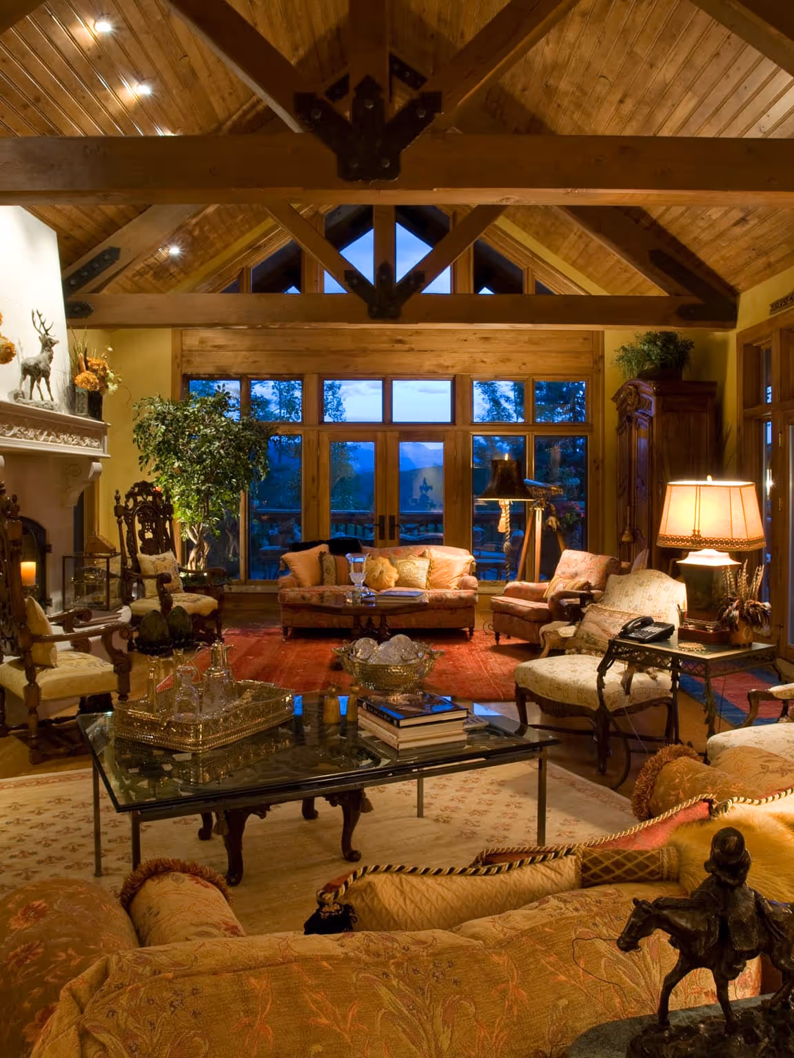 Cozy rustic living room with high wooden ceiling, large windows showing evening sky, antique furniture, and warm lighting.