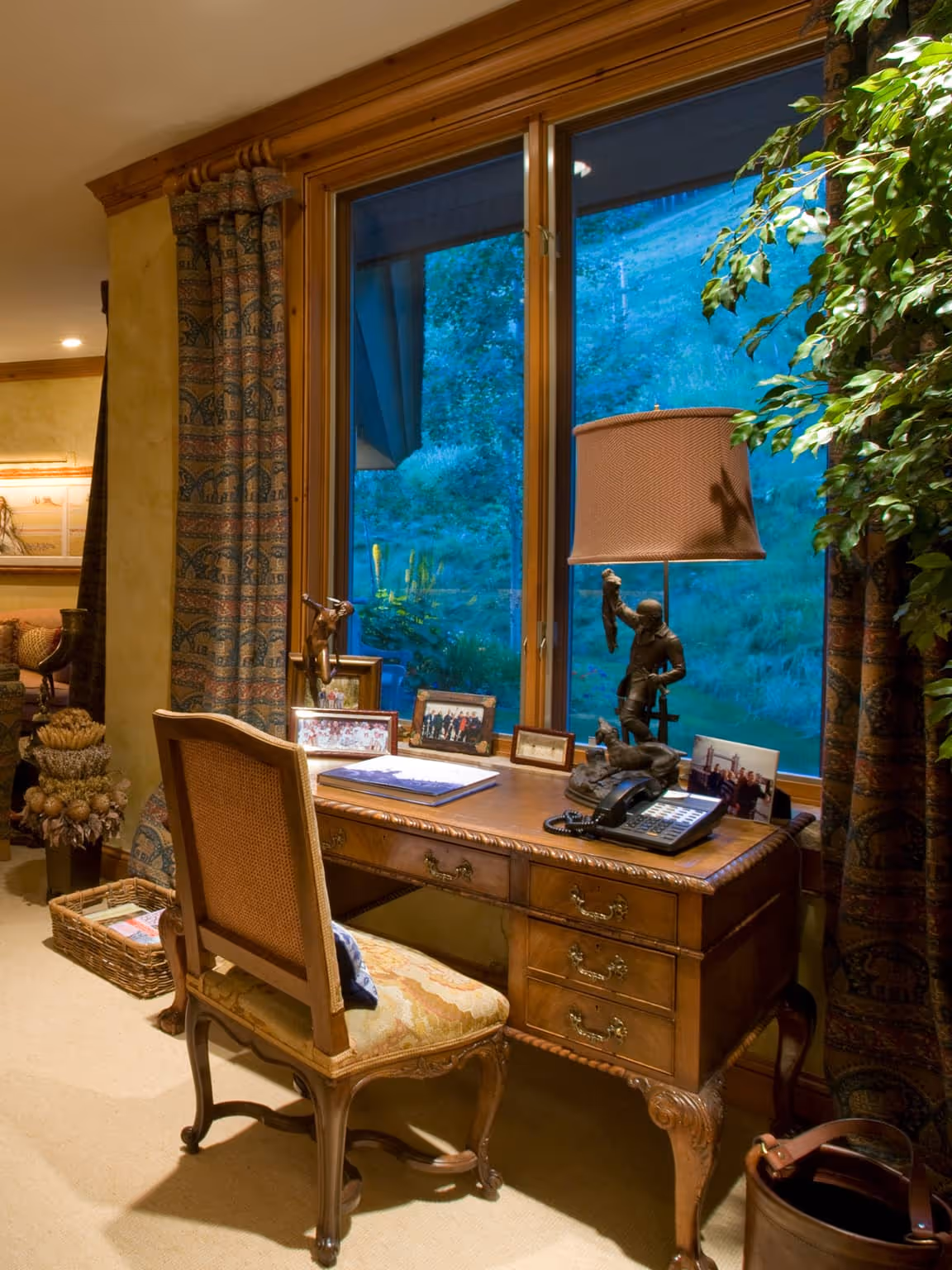 Wooden desk with vintage chair by a large window showing greenery outside, decorated with framed photos, a lamp, and sculptures.