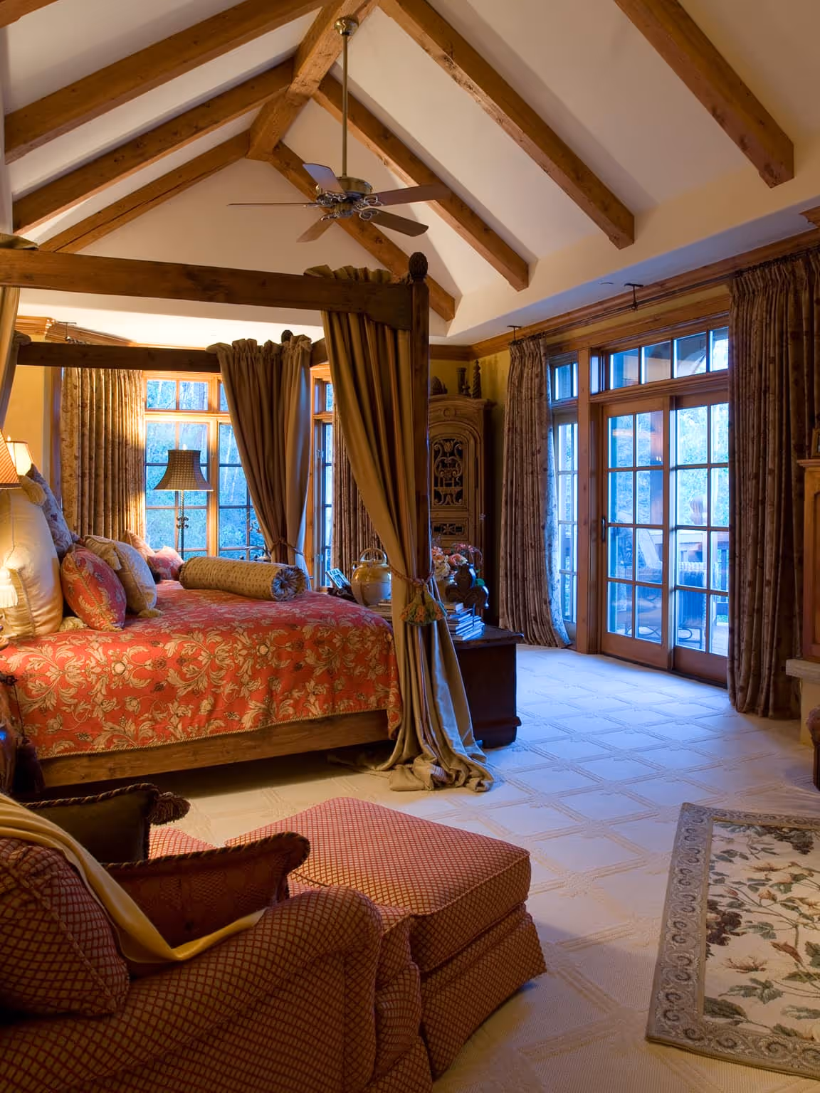 Cozy bedroom with a wooden canopy bed, patterned bedding, armchair with ottoman, ceiling fan, and large windows with curtains.
