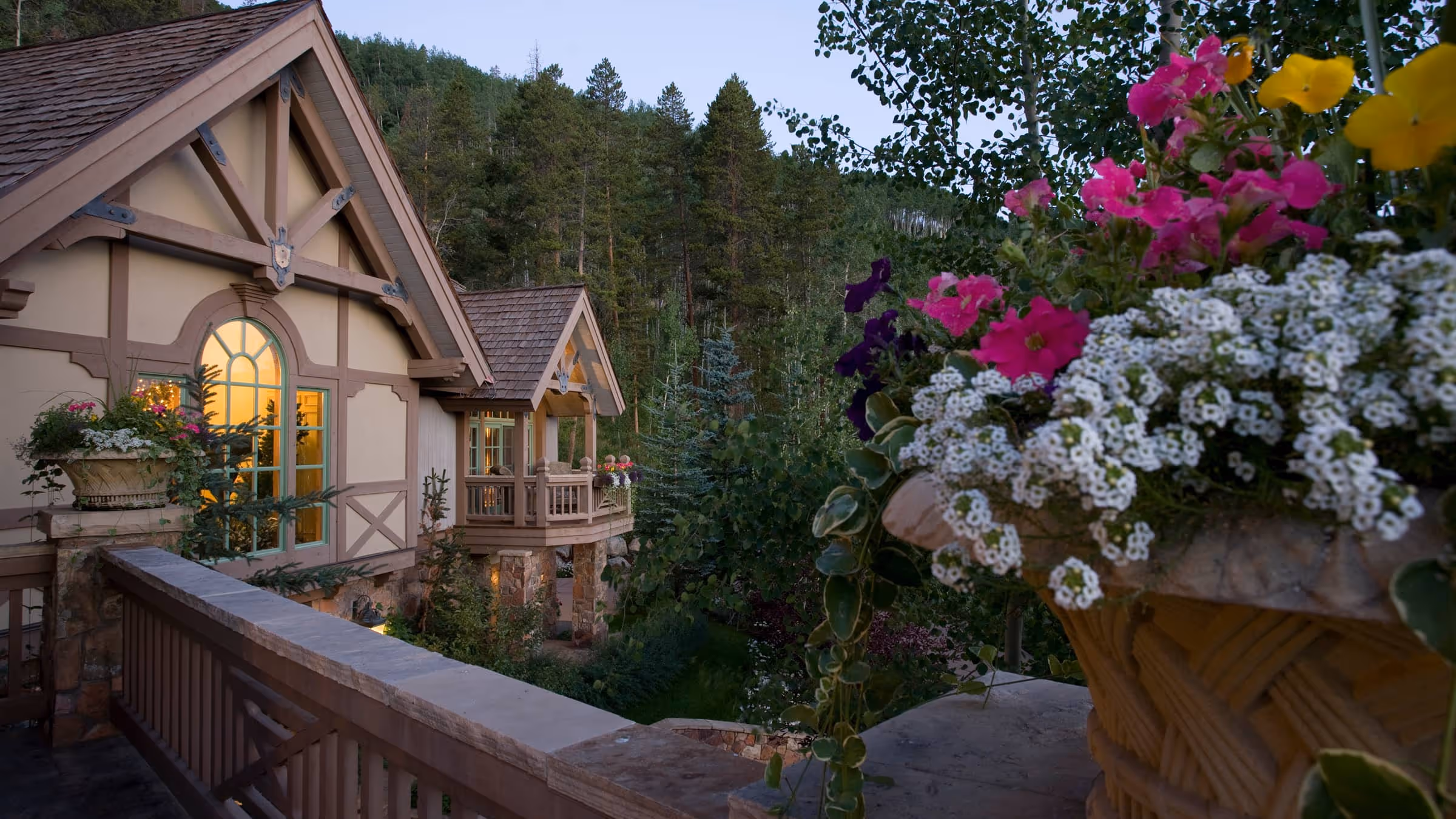 Cozy lodge with wooden beams and lit windows surrounded by pine trees and colorful flowers in planters at dusk.