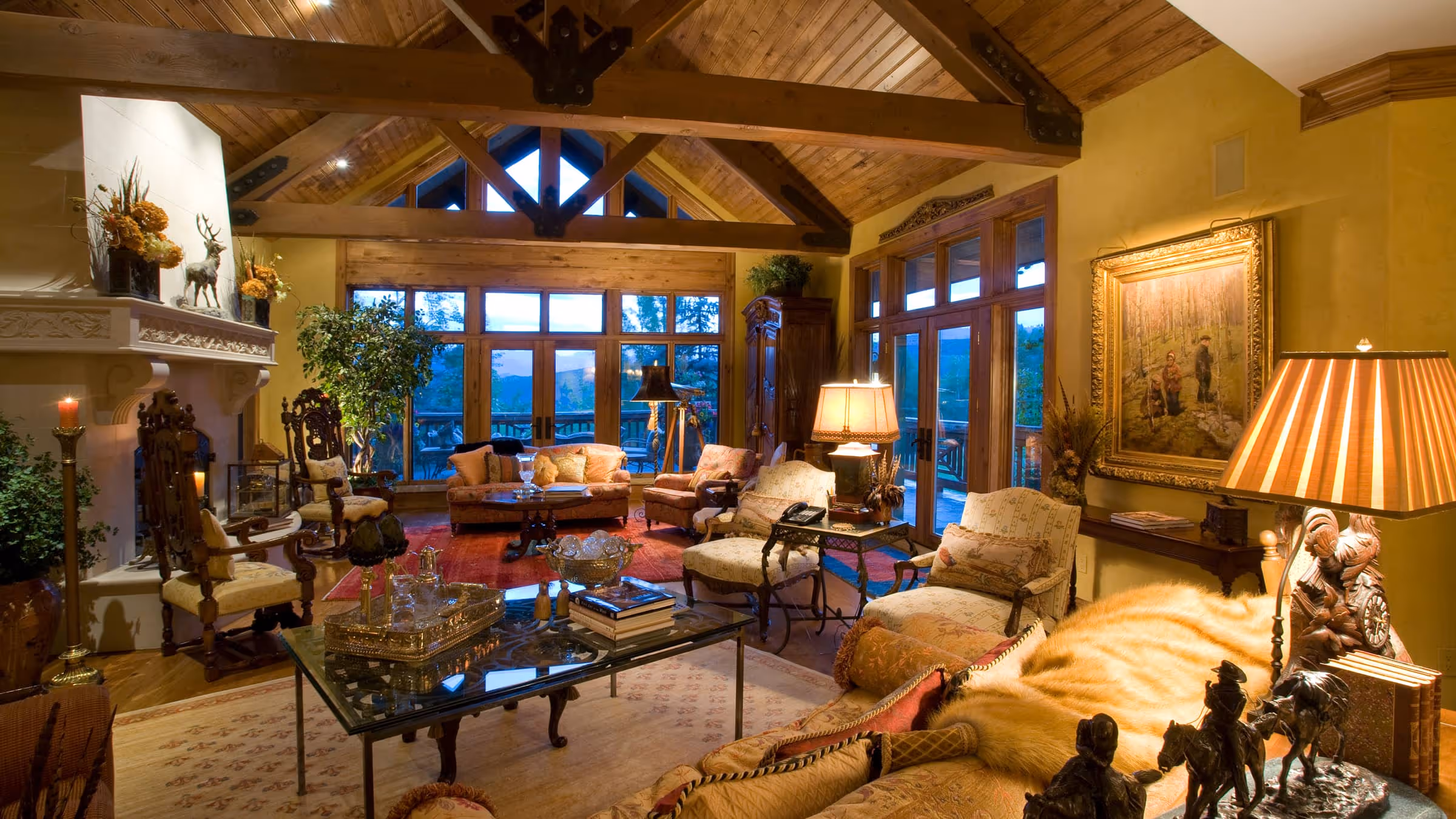 Cozy rustic living room with wooden vaulted ceiling, large windows, ornate furniture, and warm lighting.