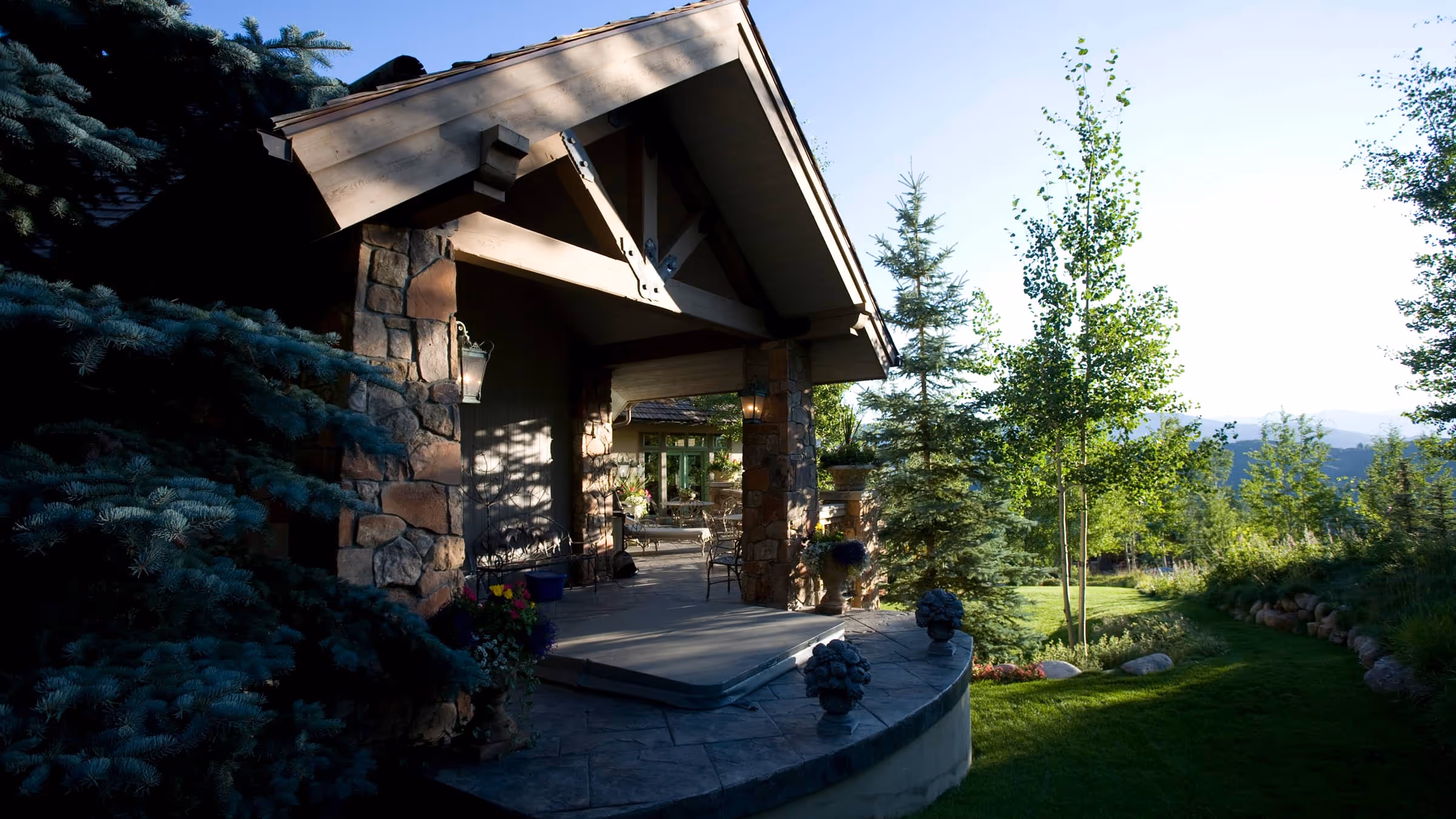 Stone and wood lodge porch with outdoor chairs surrounded by pine trees and green landscaping.