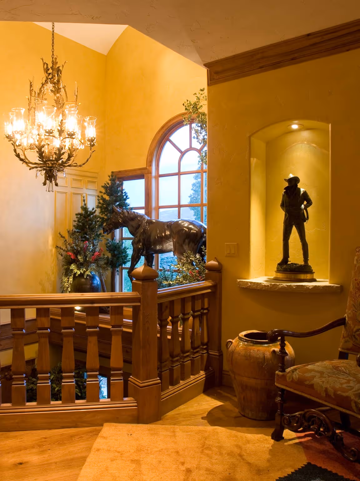 Cozy indoor space with yellow walls, wooden railing, chandelier, horse sculpture near window, cowboy statue in niche, and ornate chair.